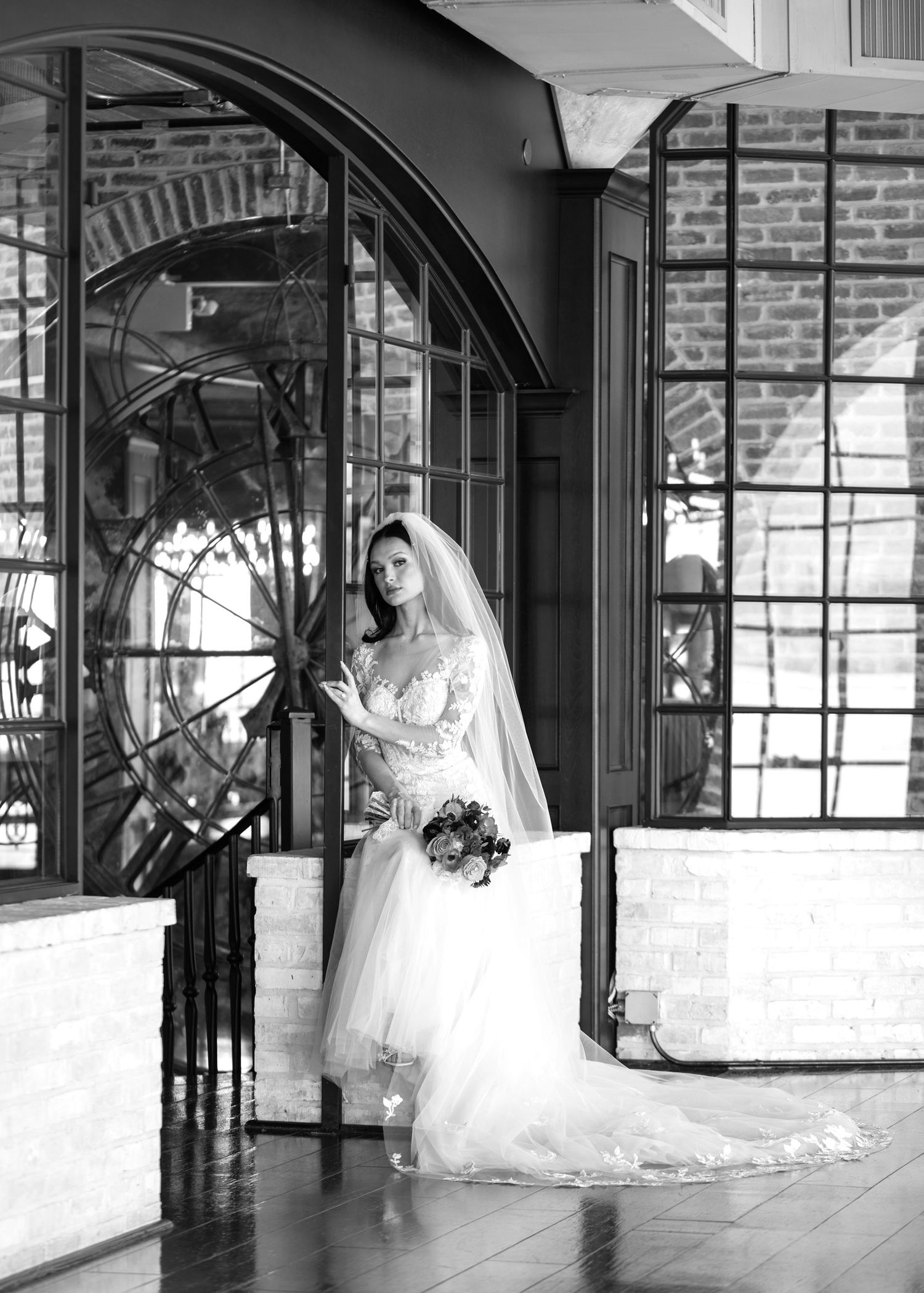 A bride in a flowing white gown stands in an elegant brick-walled venue with arched windows and reflective surfaces.