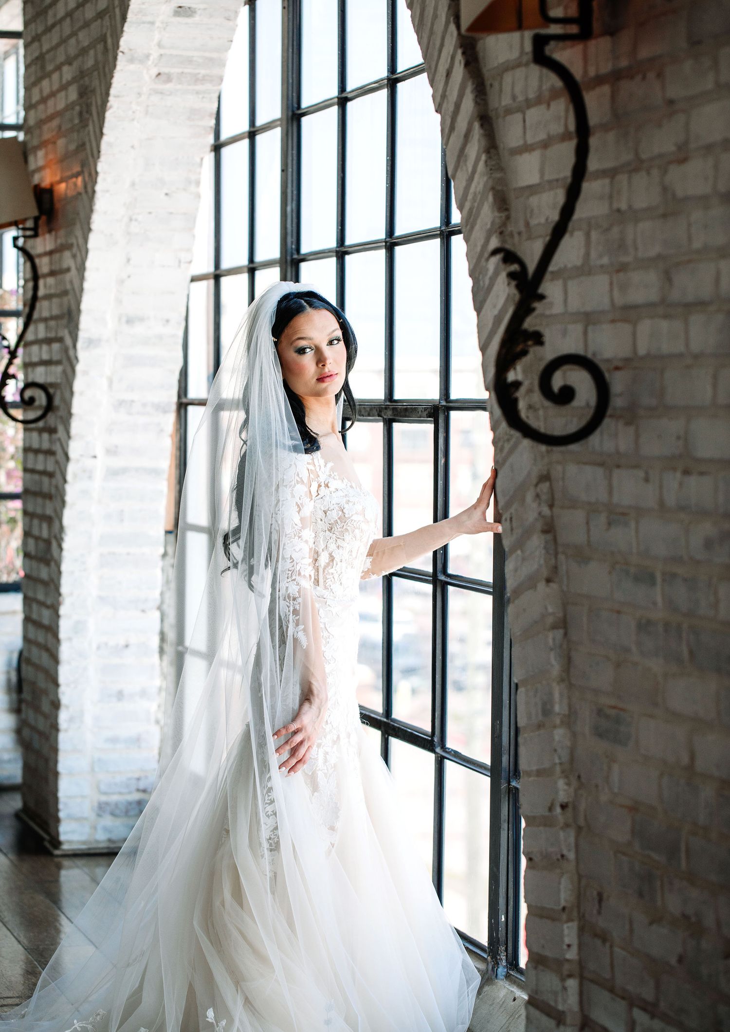 Elegant bride in white wedding dress and flowing veil poses gracefully against industrial brick wall and windows.