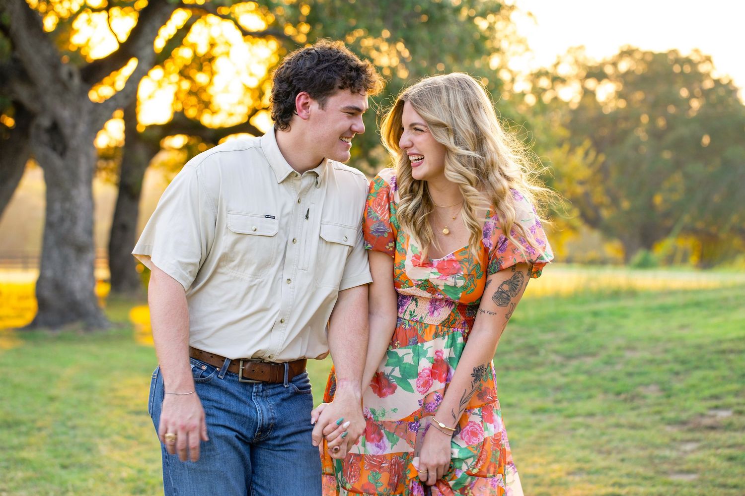 A couple shares a romantic moment at sunset in a grassy field wearing casual and floral attire.
