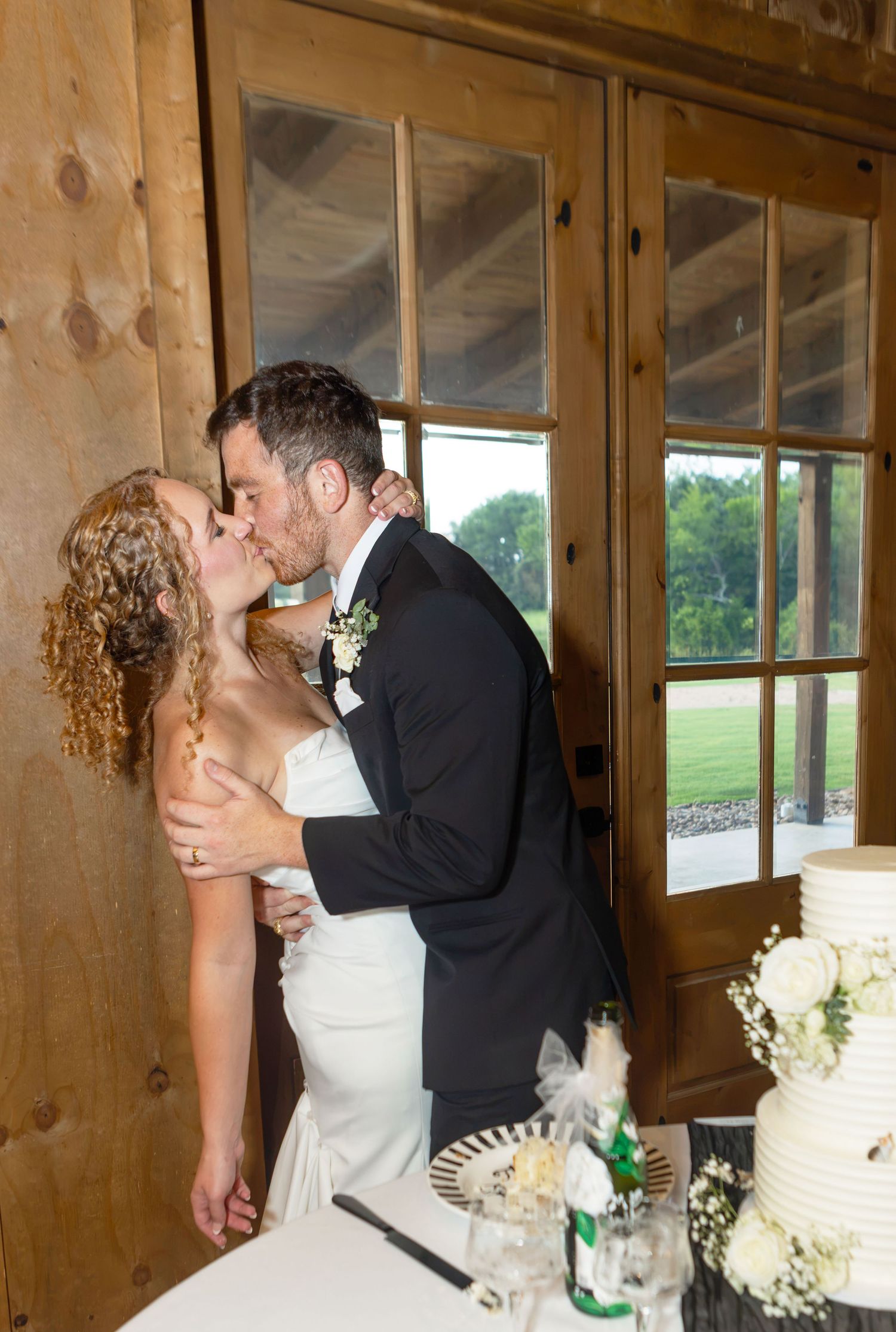 Bride and groom share a romantic kiss near wedding cake in rustic barn venue with wooden walls and glass doors.