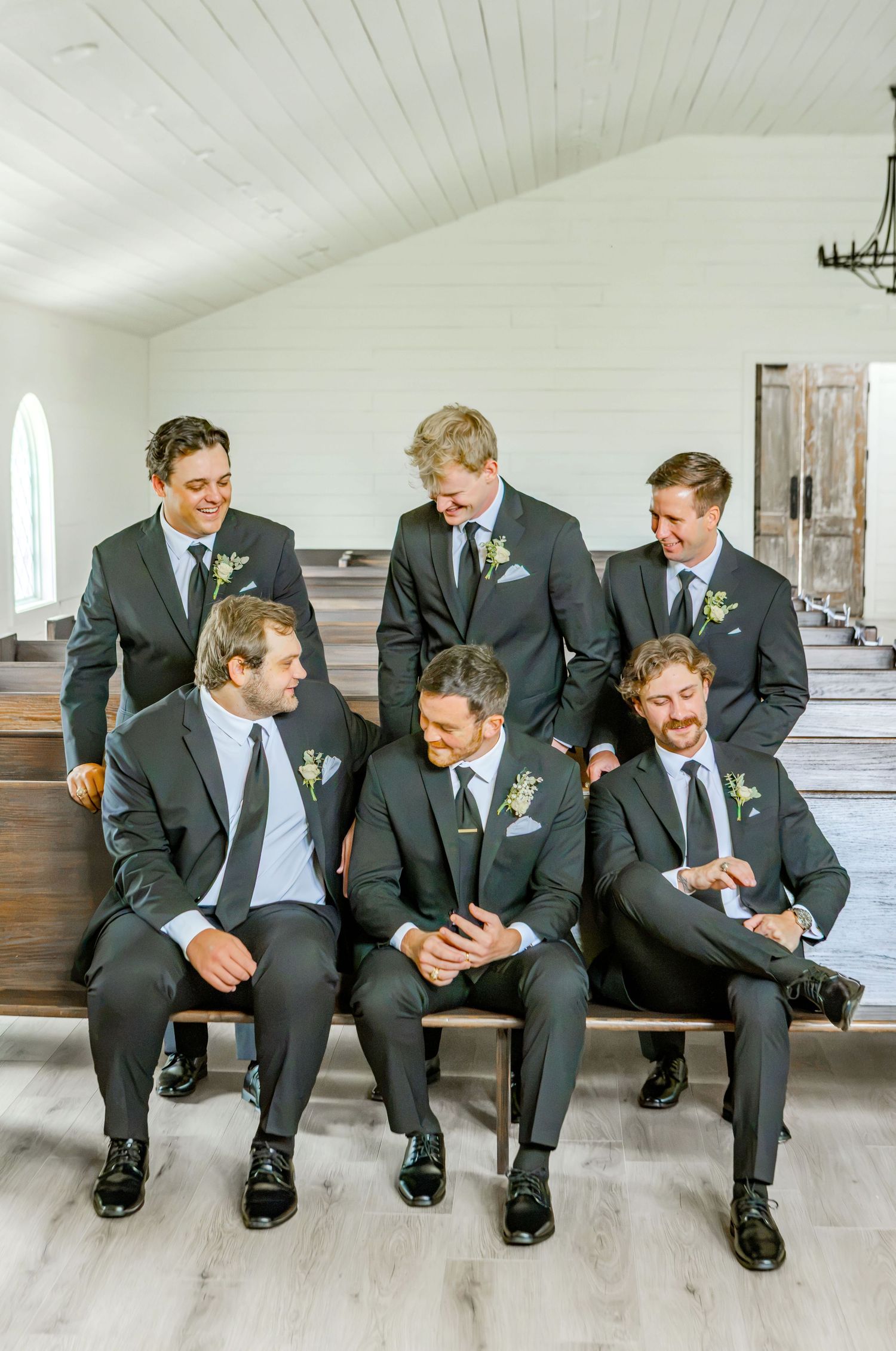 Seven groomsmen in dark suits with boutonnieres pose on wooden church pews, laughing together at a wedding.