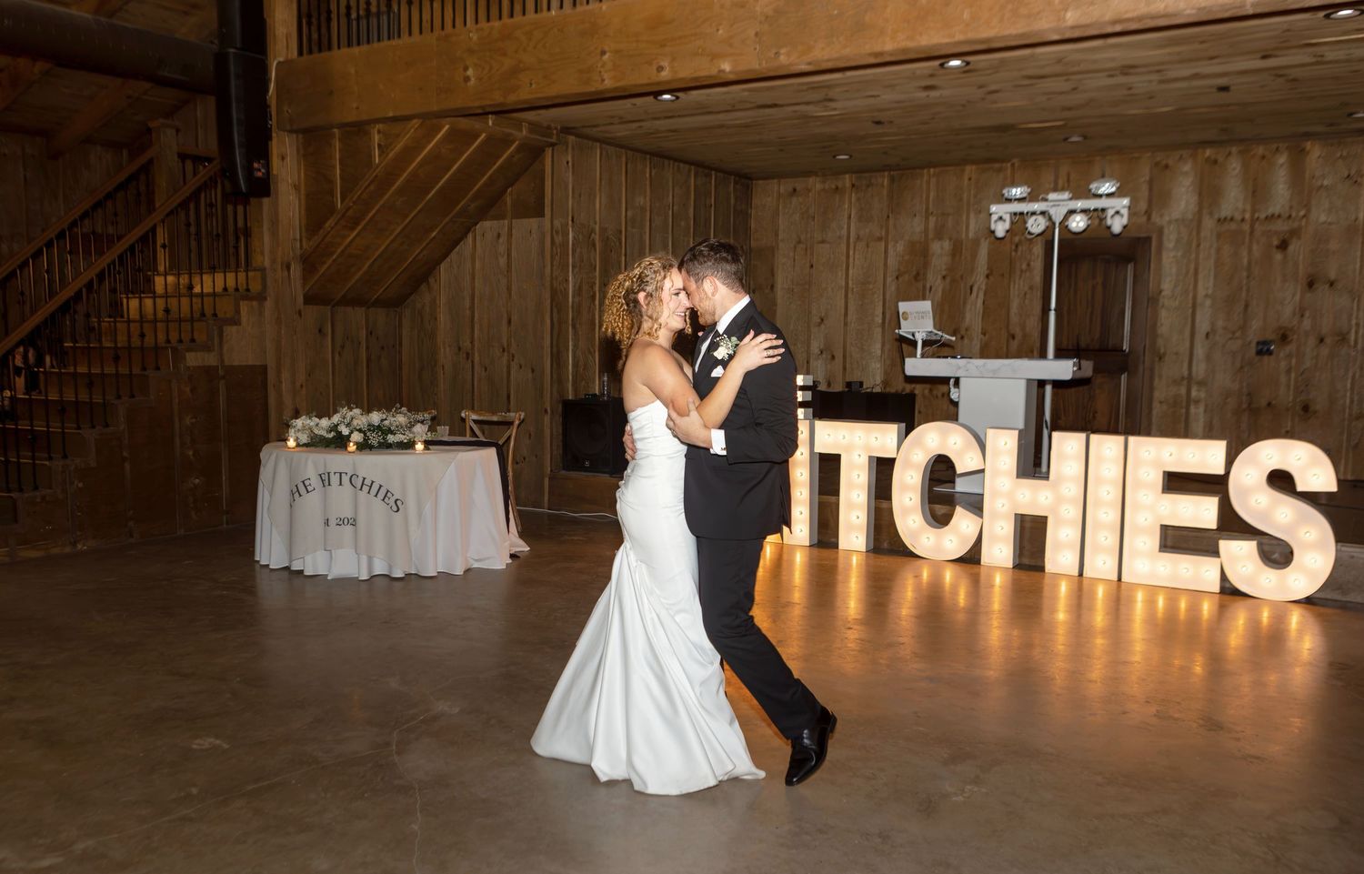 Bride and groom share first dance in rustic barn venue with illuminated marquee letters spelling ITCHIES in background.