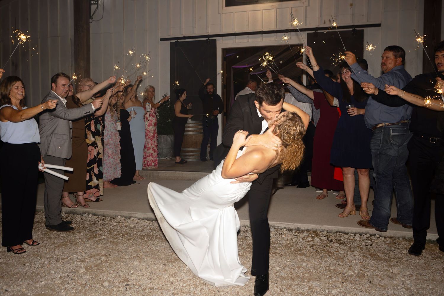 Bride and groom share romantic dip kiss surrounded by guests holding sparklers at outdoor wedding reception.