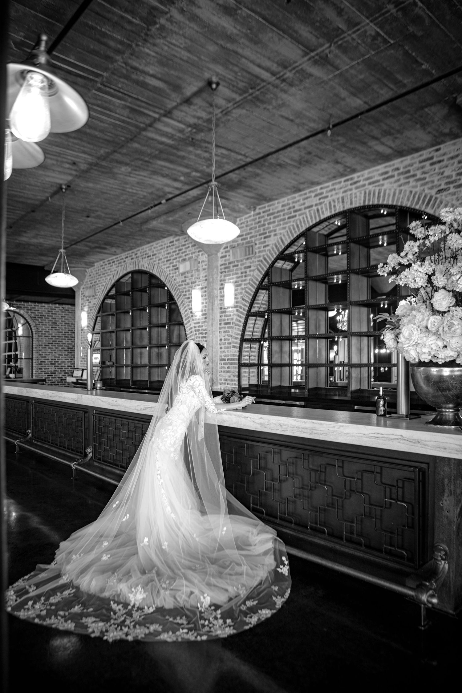 A bride in a flowing white gown and veil stands in an elegant venue with arched brick walls and pendant lighting.