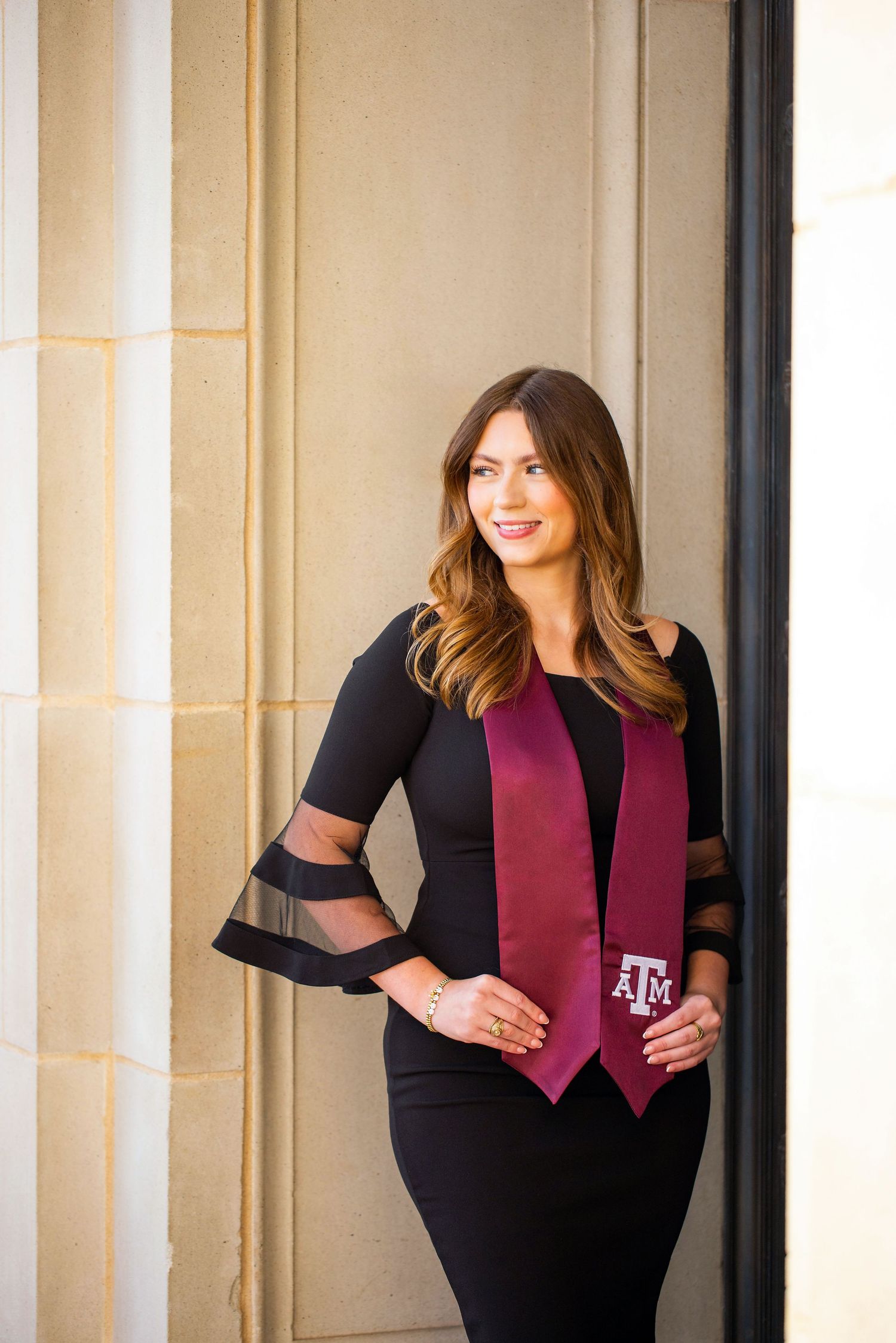 A smiling woman with long brown hair wearing a black and burgundy outfit stands in front of a light-colored wall.