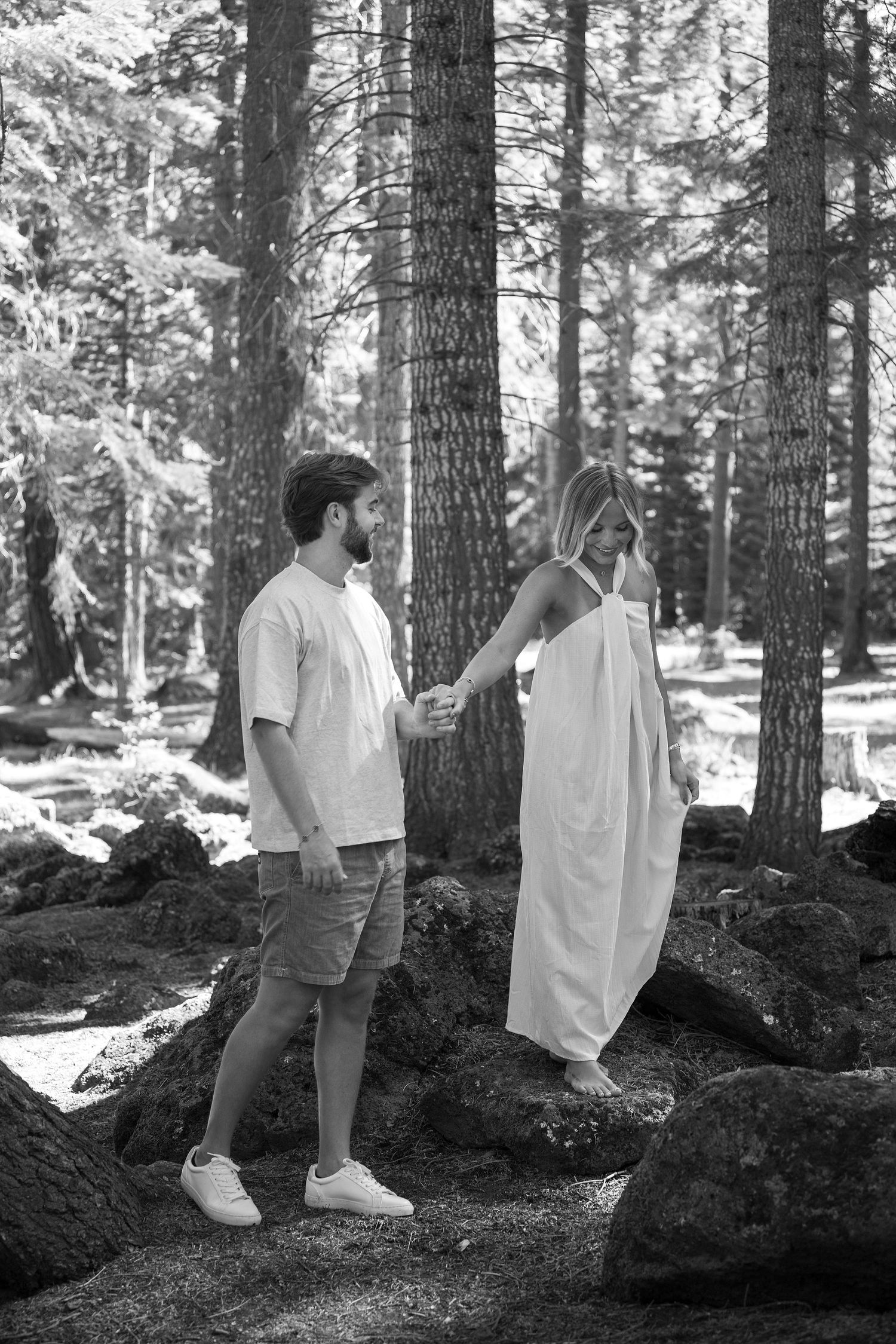 Black and white photo of a couple holding hands while walking on rocks in a pine forest, woman in white dress.