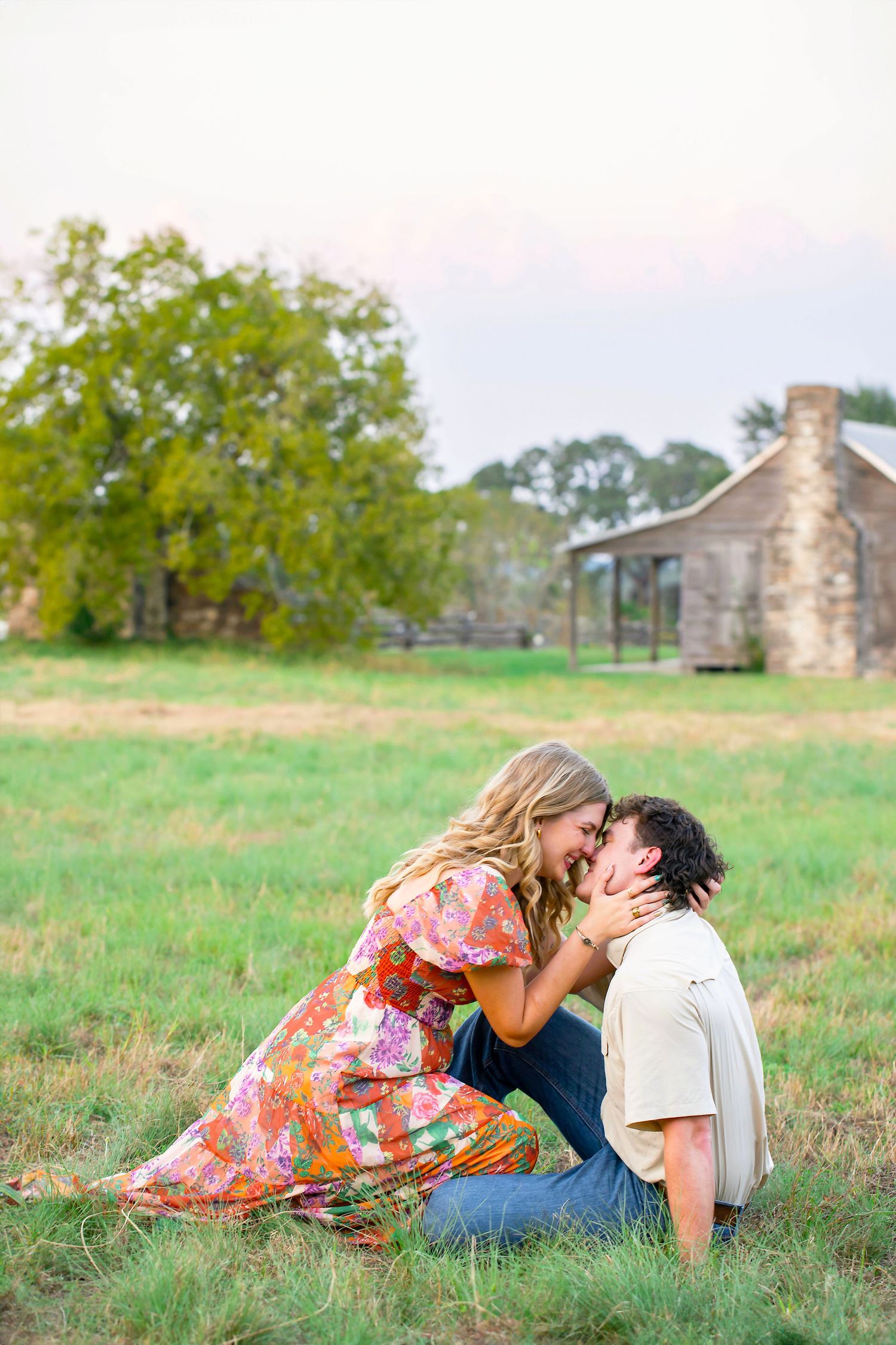 Couple in floral dress and casual wear lying together on grassy field with rustic stone building in background.
