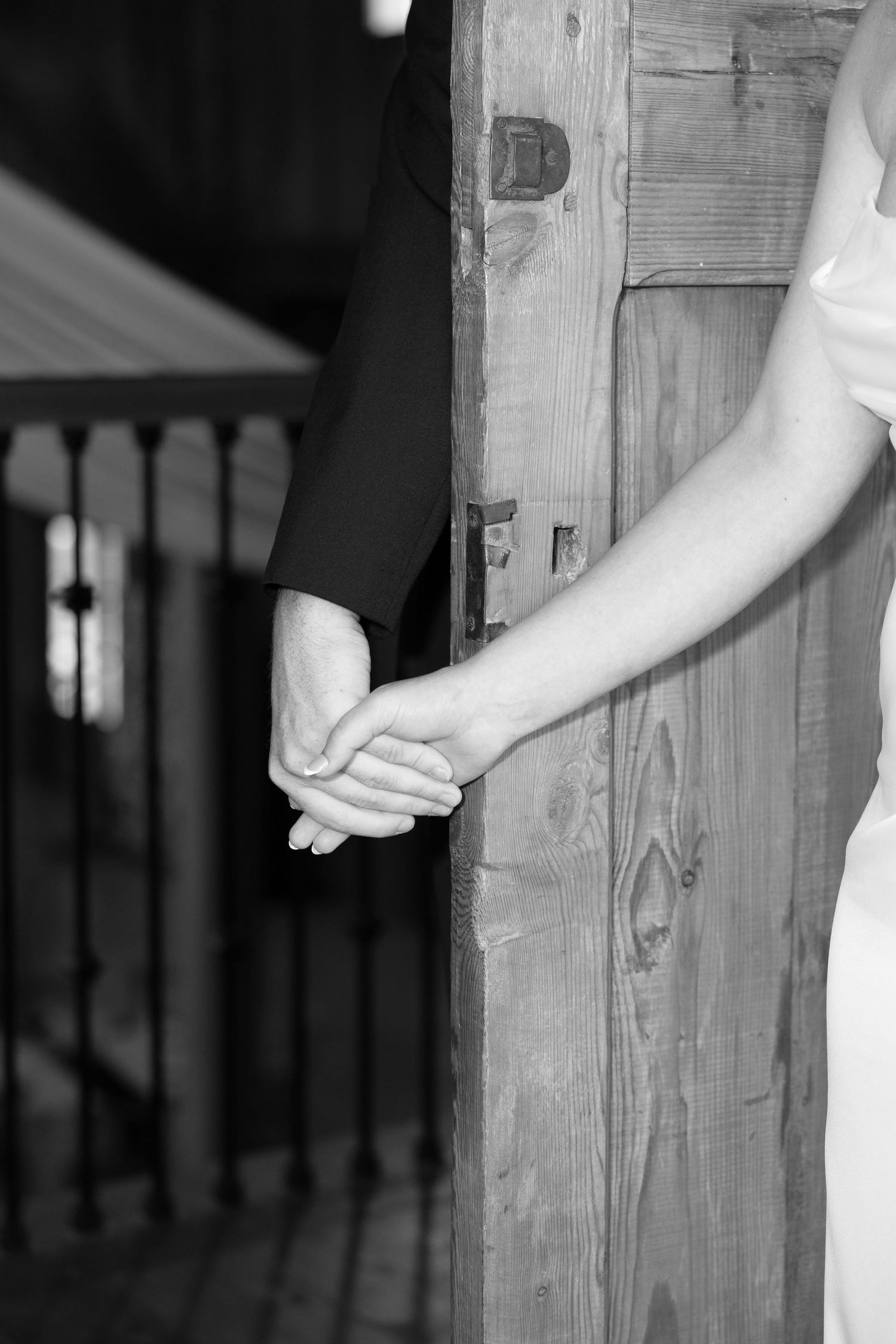 Black and white photo of a bride and groom holding hands beside a rustic wooden door at their wedding.