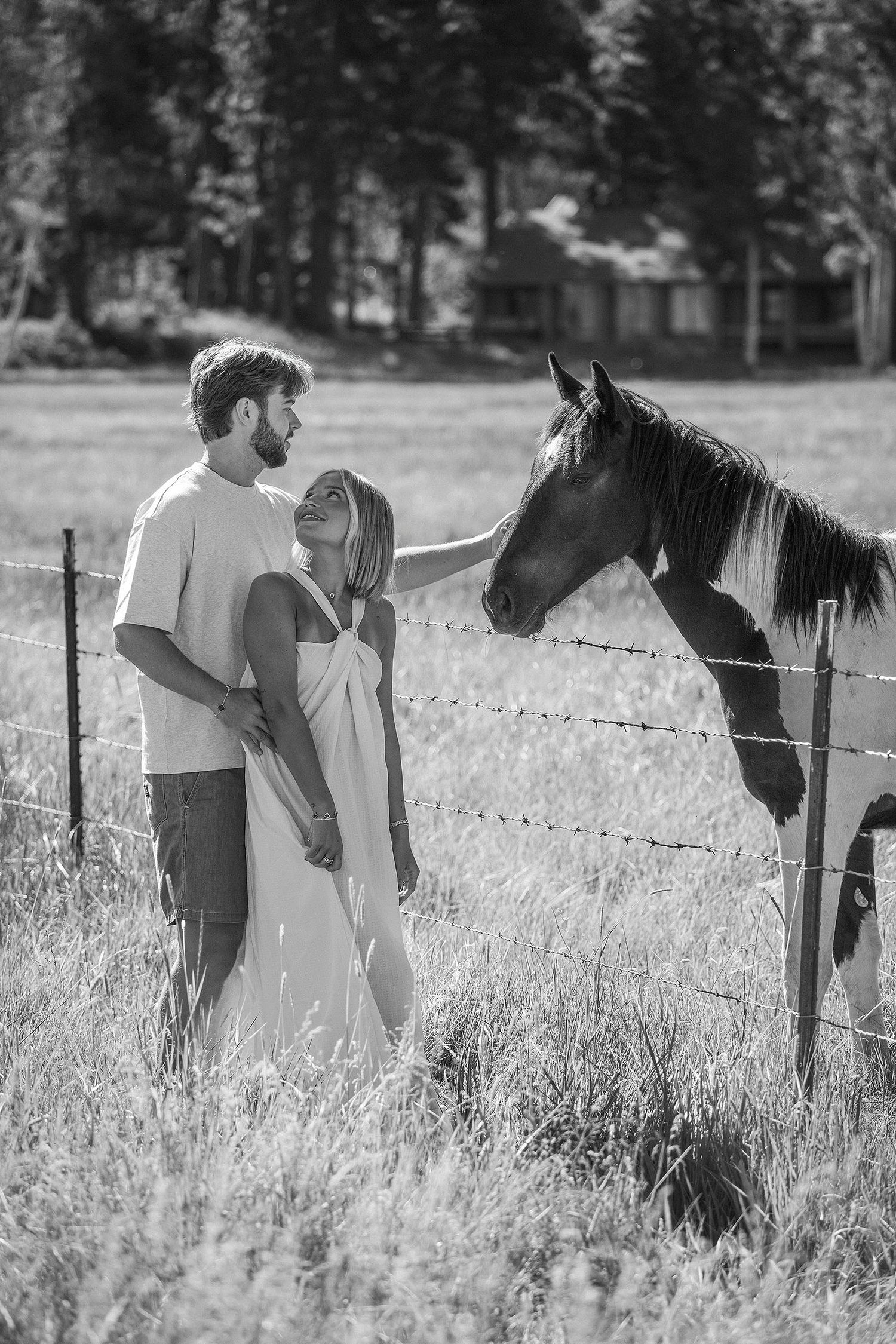 Black and white photo of a couple standing by a barbed wire fence petting a horse in a rural field.