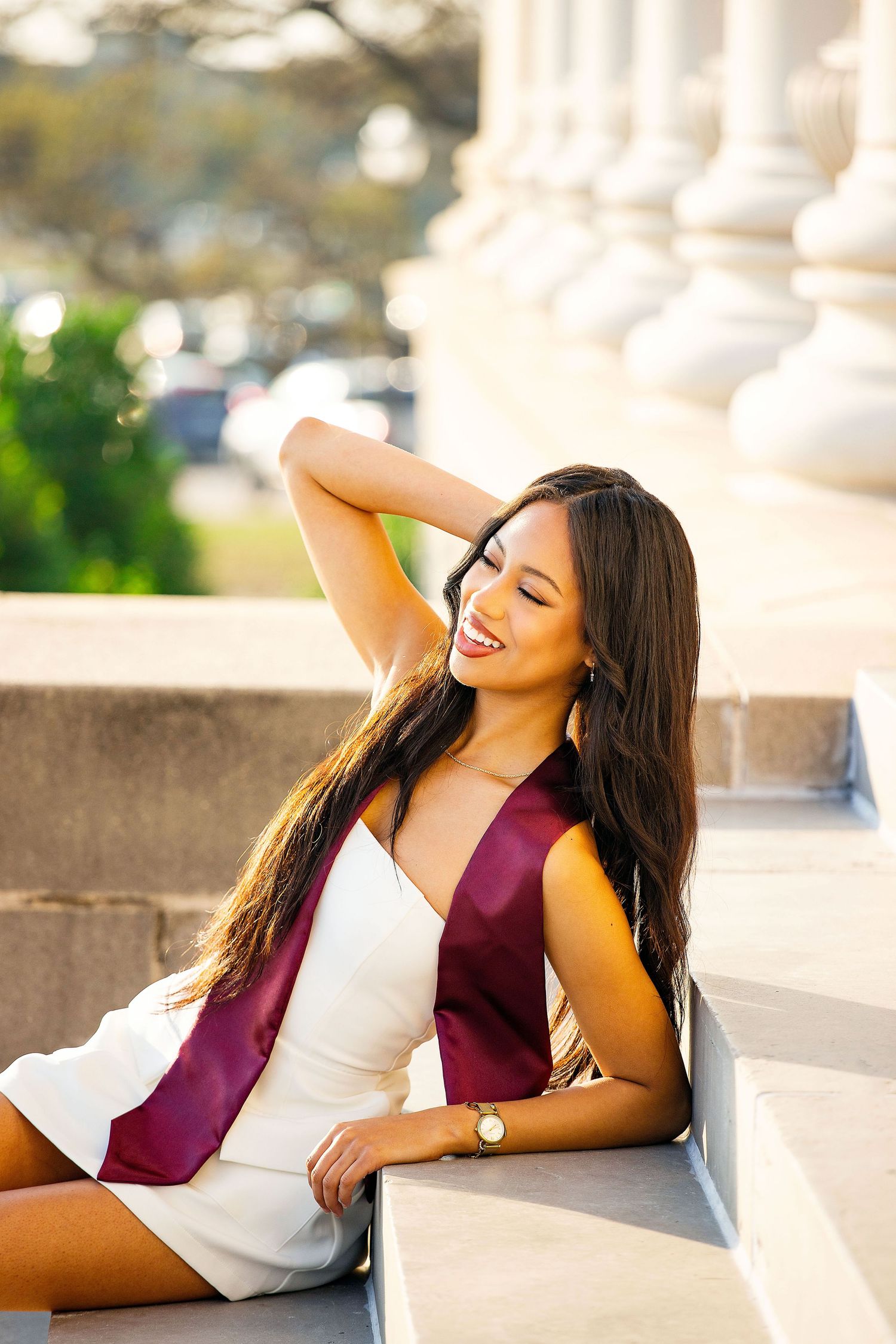 A graduate in a white dress and maroon graduation stole sits on stone steps while smiling and posing for a portrait.