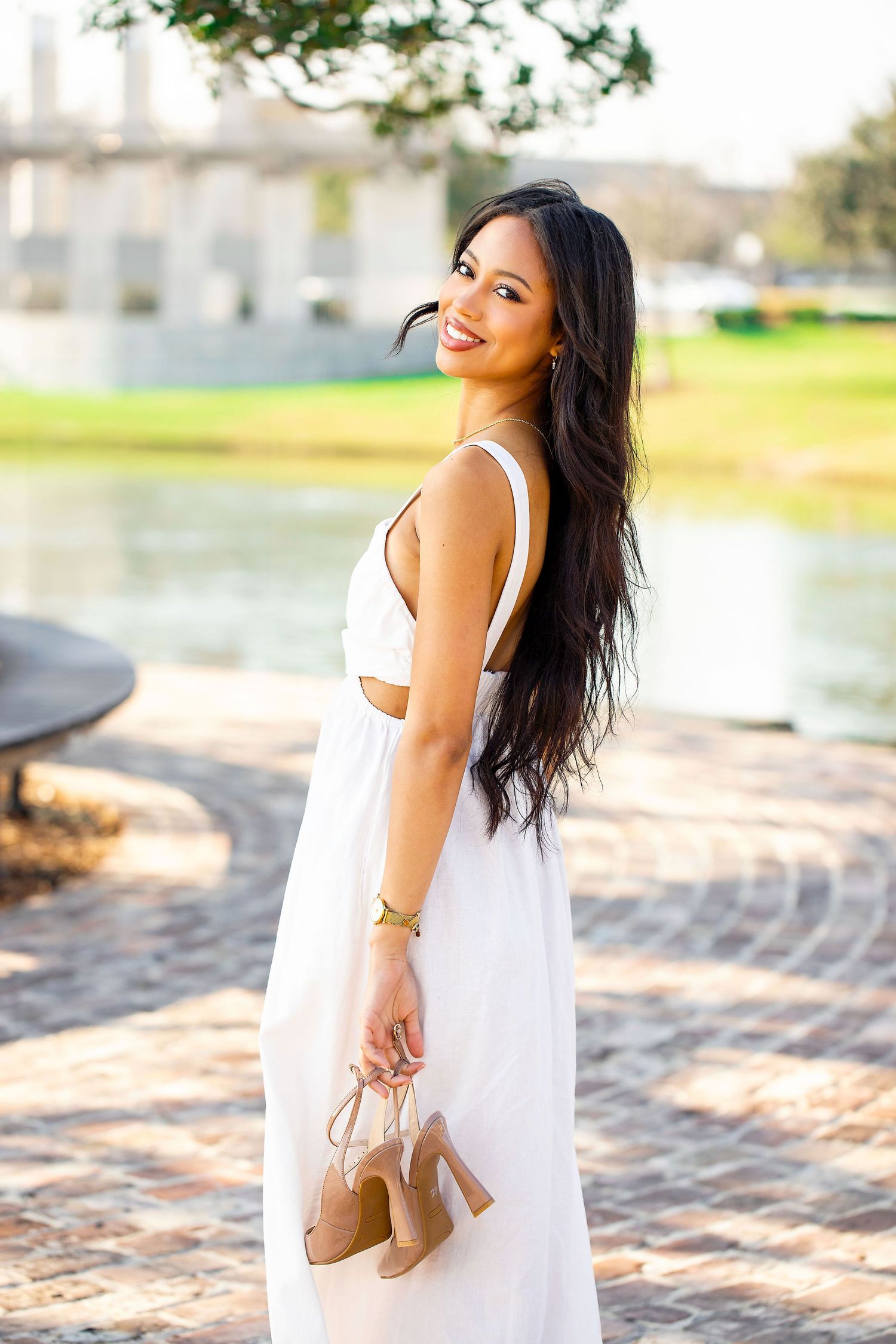 A white maxi dress outfit photographed by a lake with tan heels being held in hand on a sunny day.