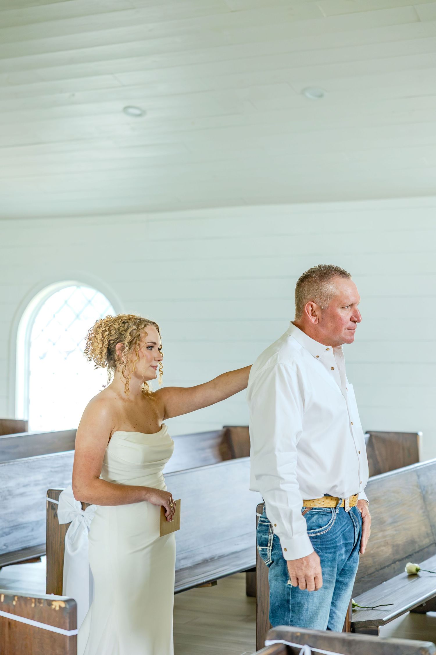 Bride in white strapless gown touches groom's shoulder in a white chapel with wooden pews during wedding first look.
