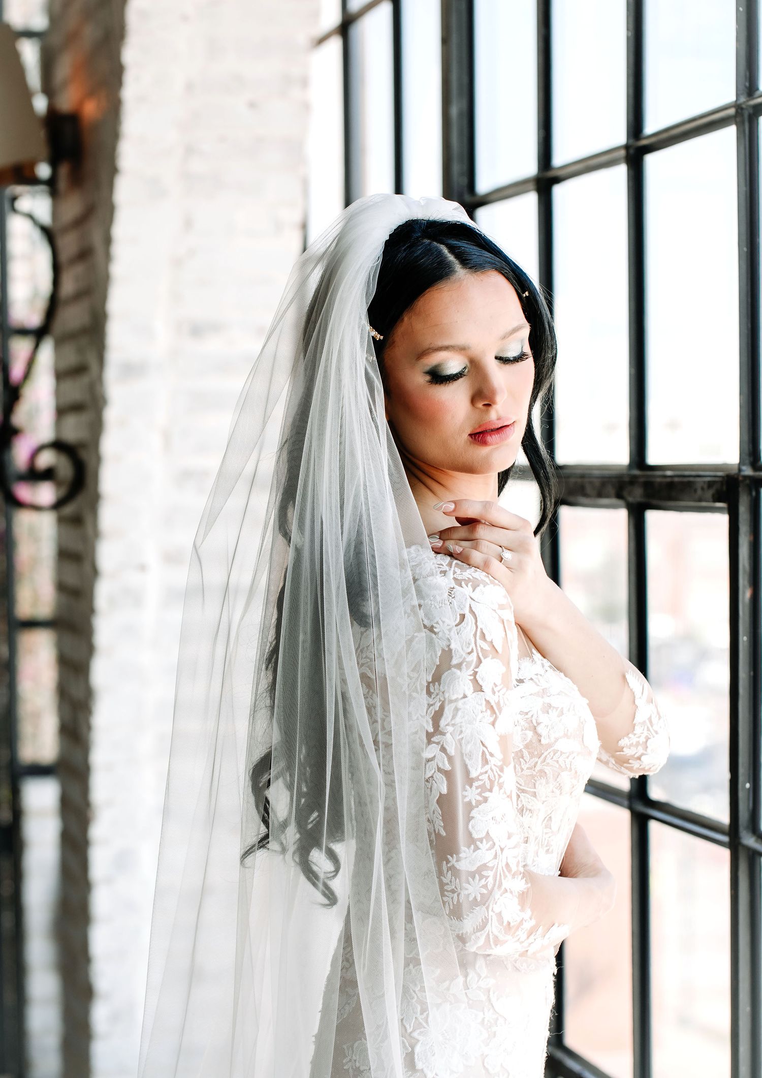A bride in a white lace wedding dress and veil poses elegantly by large industrial windows in an artistic portrait.
