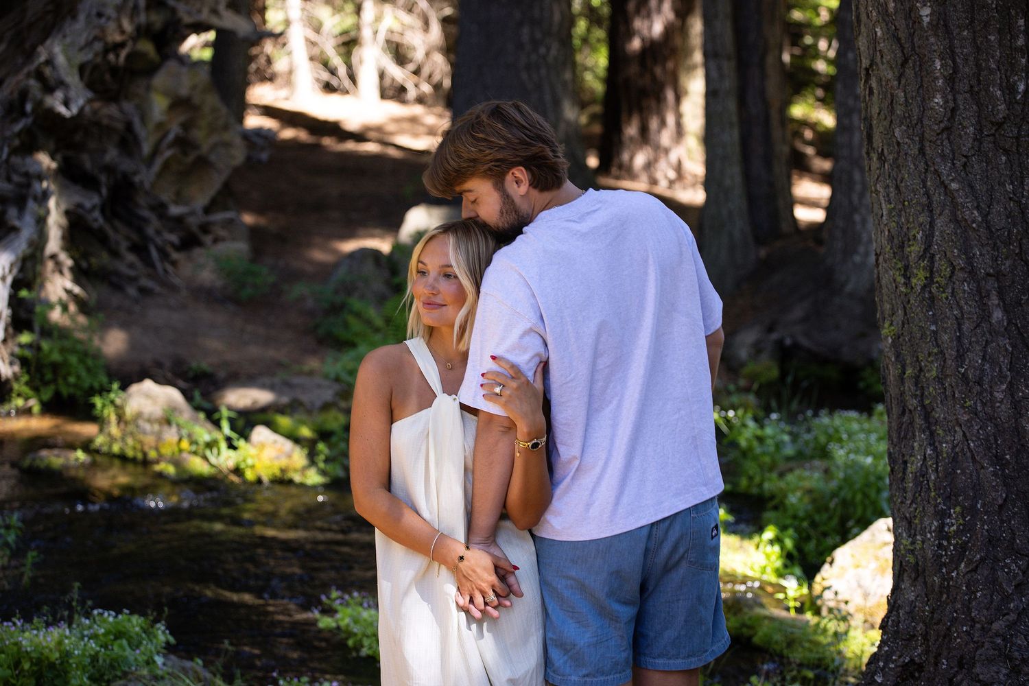 A couple in a forest setting, woman in white dress and man in blue shorts embracing among tall trees.