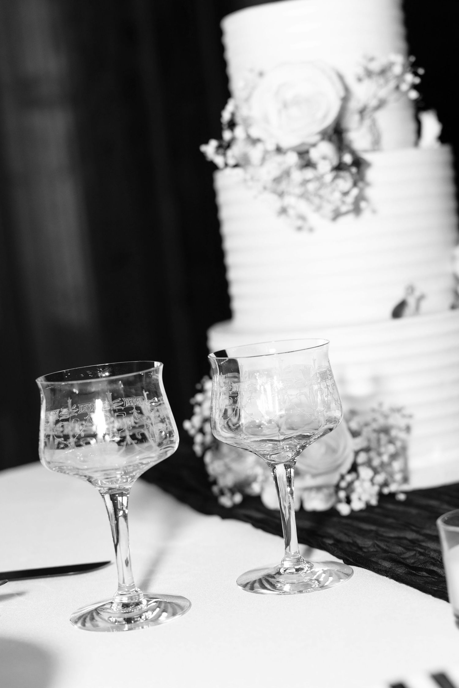 Two elegant crystal glasses beside a tiered white wedding cake decorated with flowers, in black and white.