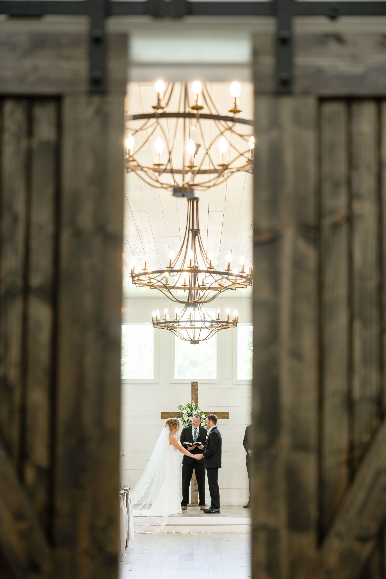 Bride and groom exchange vows beneath elegant chandeliers, framed by rustic barn doors with a wooden cross backdrop.