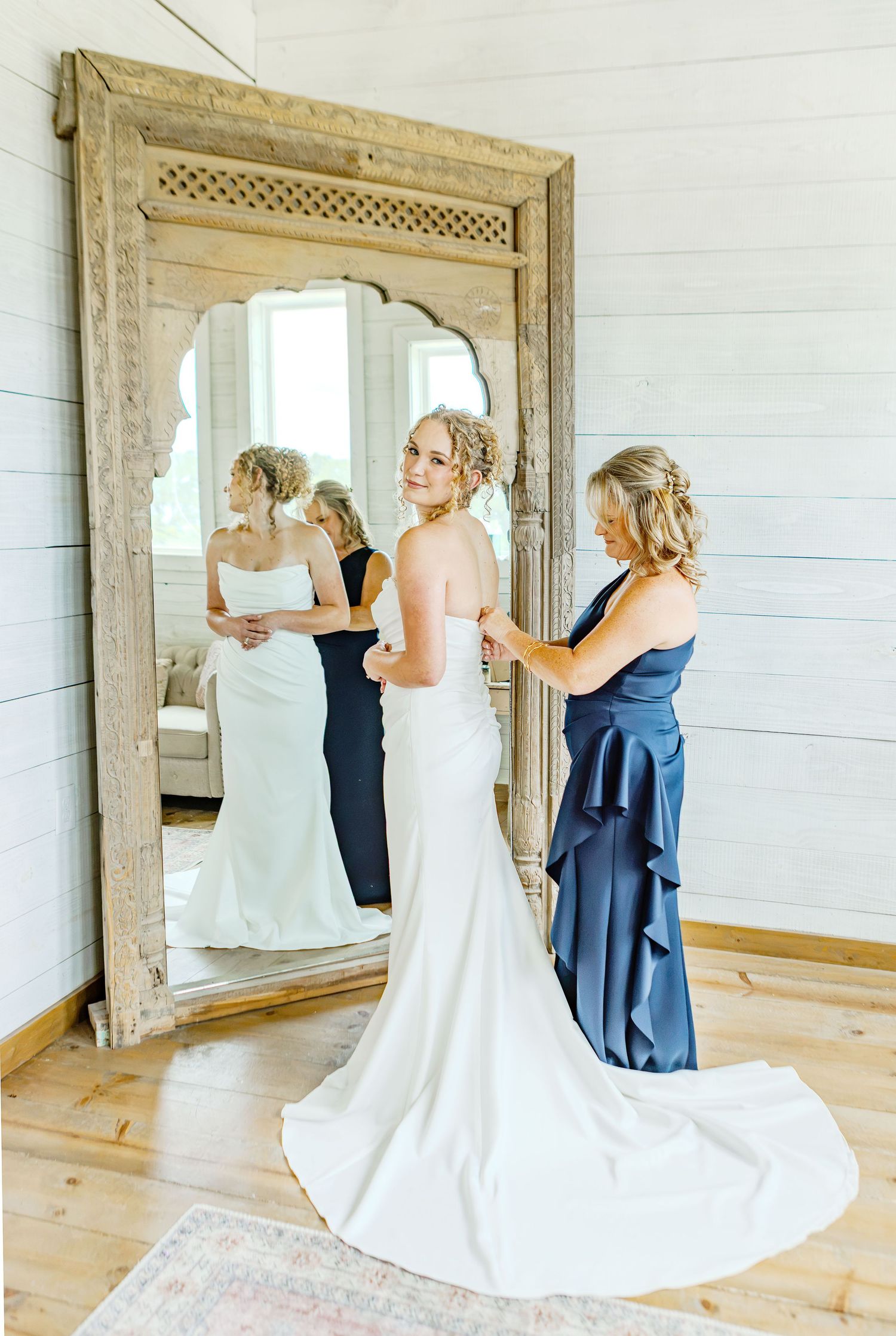 Bride in white gown getting dress fastened by bridesmaid in navy blue before large ornate mirror in bright room.