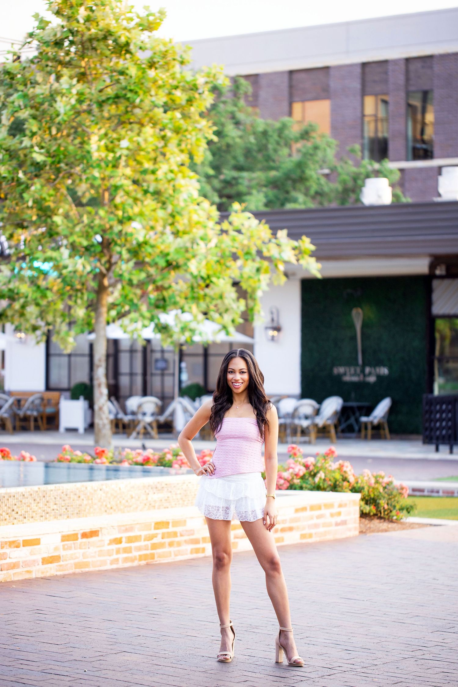 Texas A&M University graduate in pink top and white skirt  photographed in an urban setting with brick walls and trees.