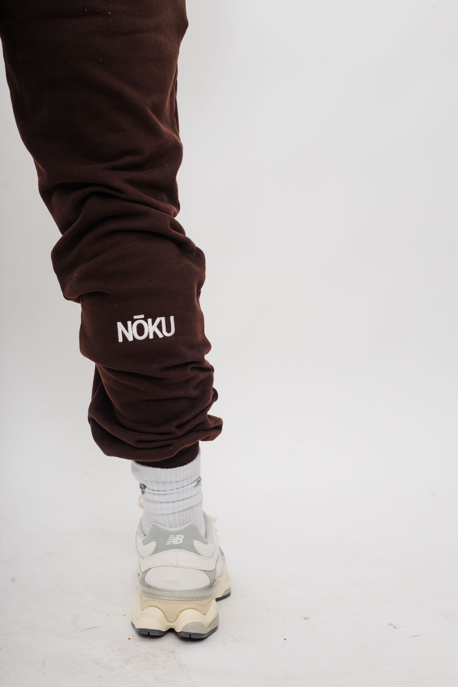 Brown sweatpants with NOKU branding and white sneakers shown against white background.