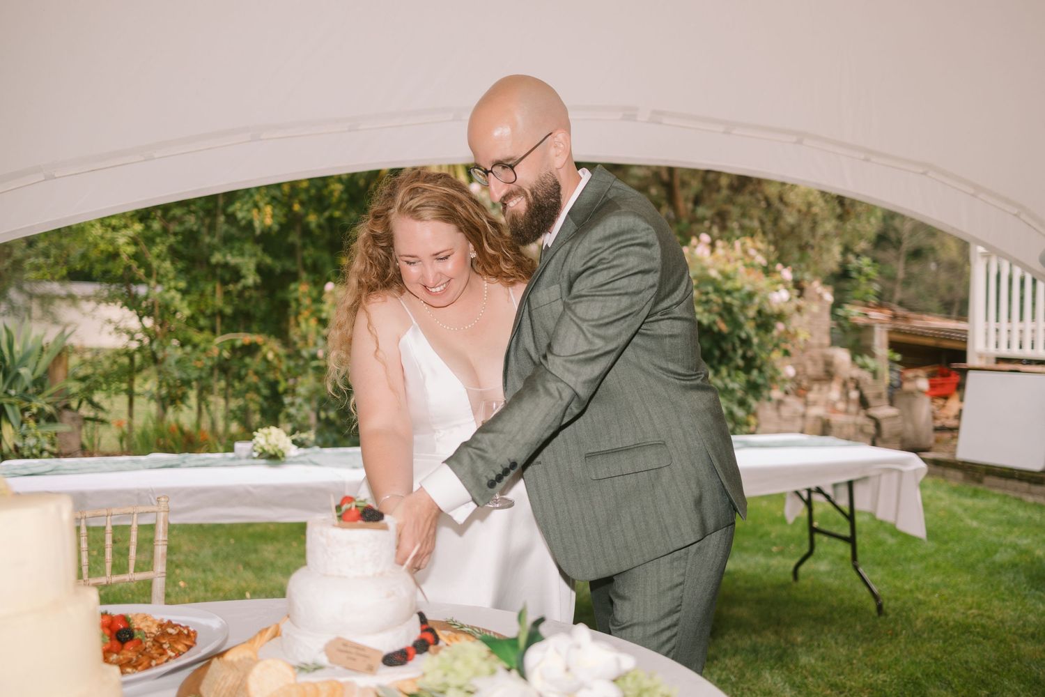 Couple cutting wedding cake together at outdoor reception under white arch.
