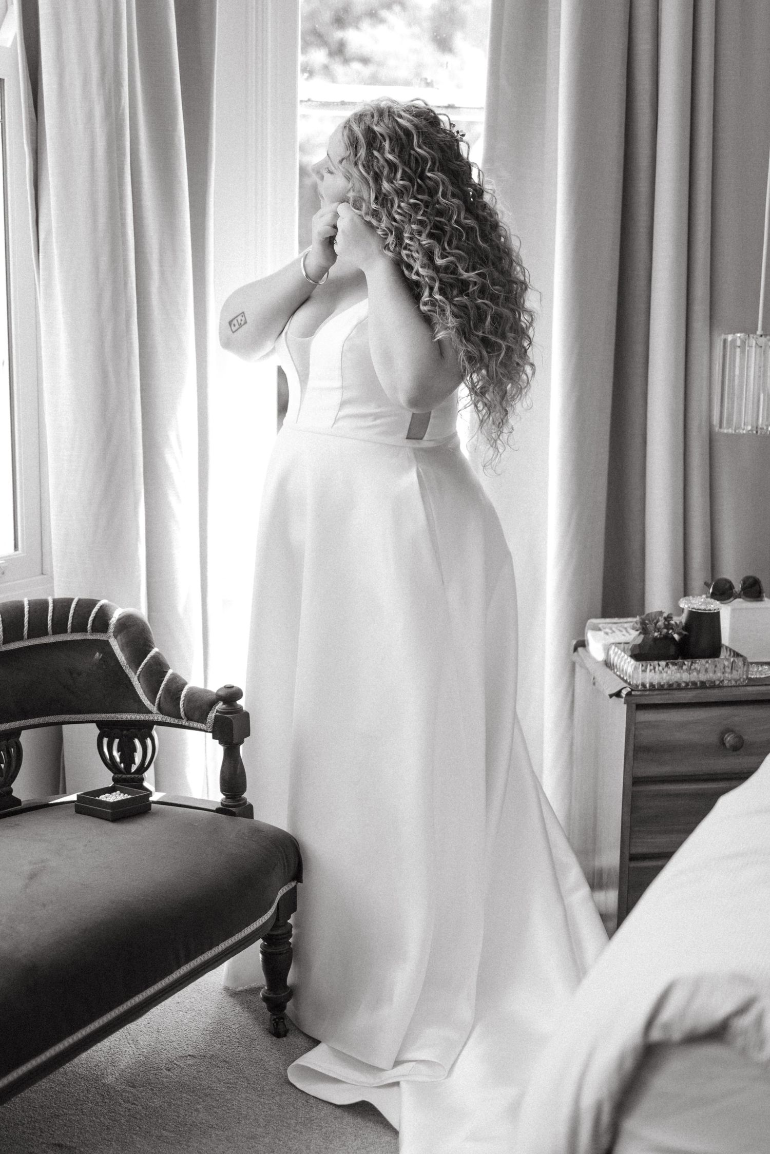 A black and white photo of a bride in a flowing white wedding dress with long curly hair gazing out a window.