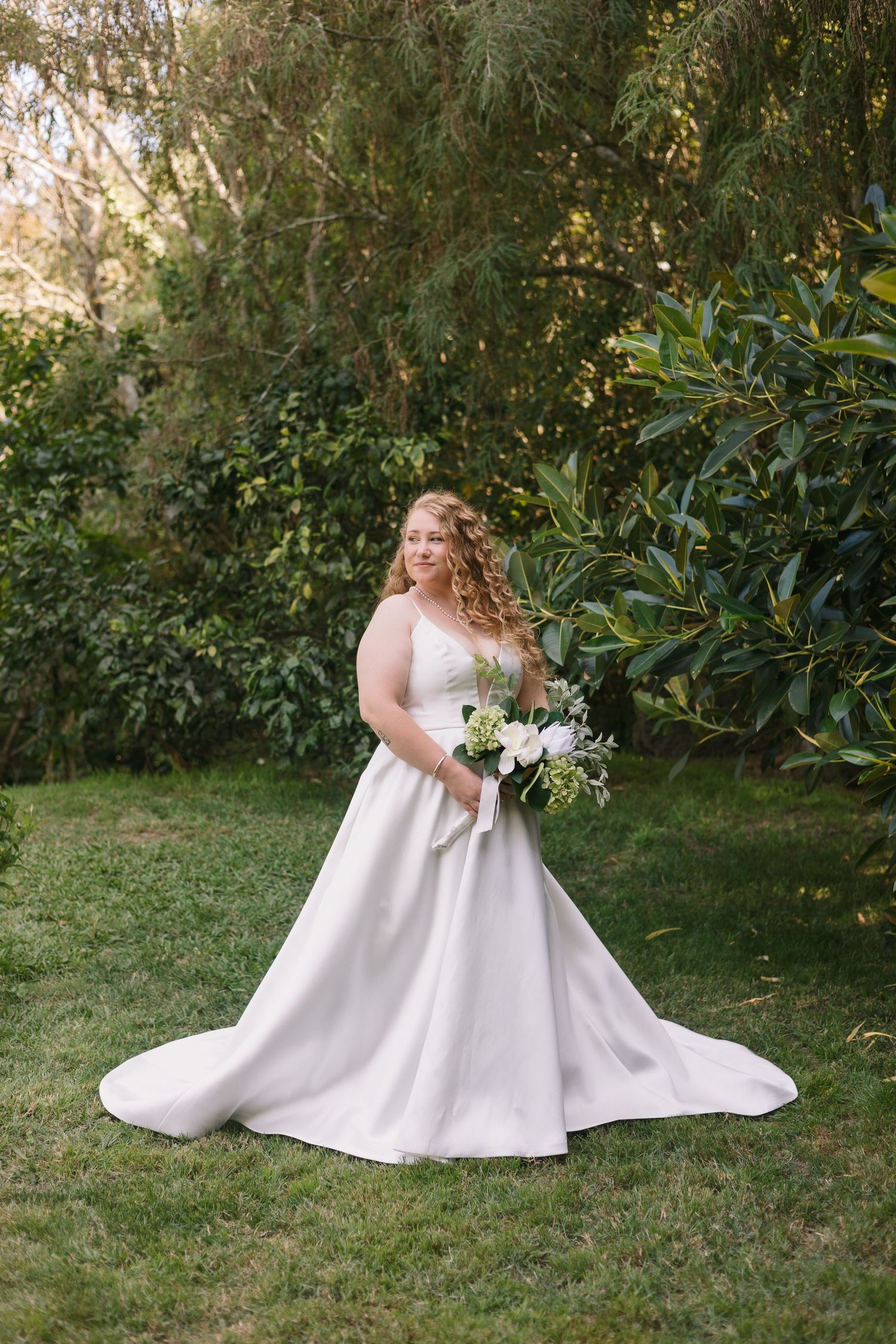 A bride in a classic white ballgown poses elegantly in a garden setting with lush greenery and holds a white floral bouquet.