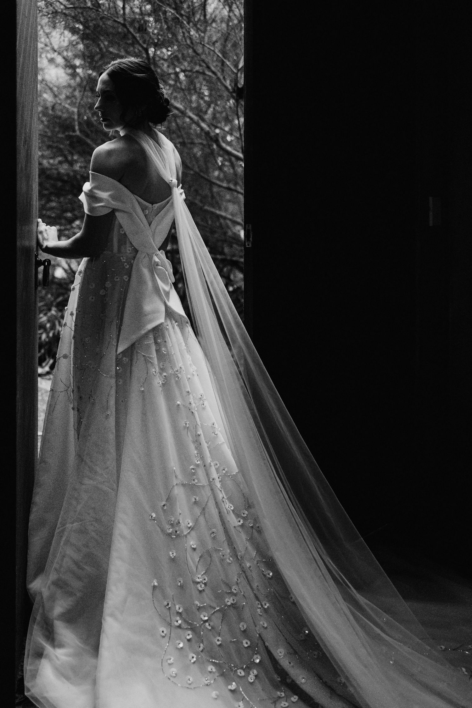 A black and white photograph shows an elegant wedding dress with floral details and a long flowing veil.