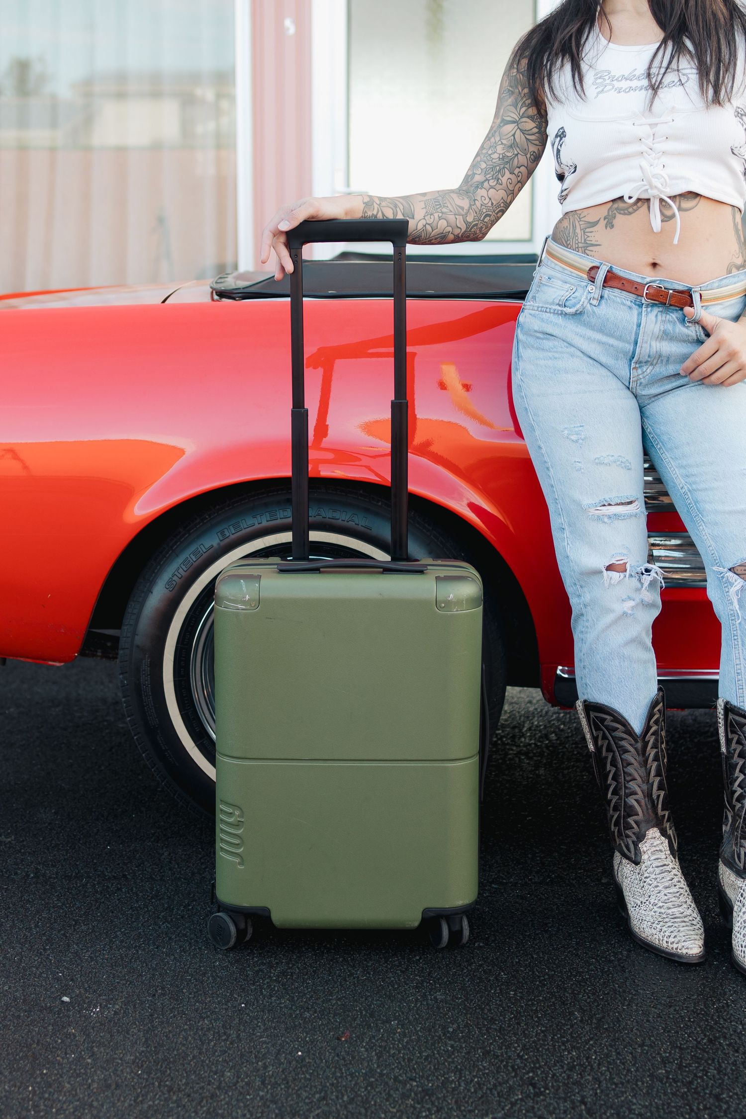 Green luggage next to a red vintage car with someone wearing ripped jeans and boots.