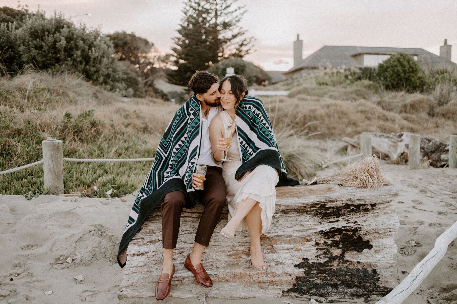 Couple sharing intimate moment on sandy beach with coastal vegetation in background.
