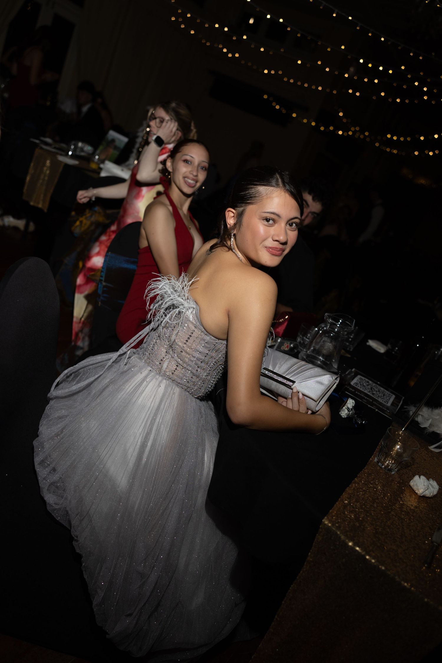 Young woman in silver feathered gown holding clutch at formal event with string lights and guests in background.