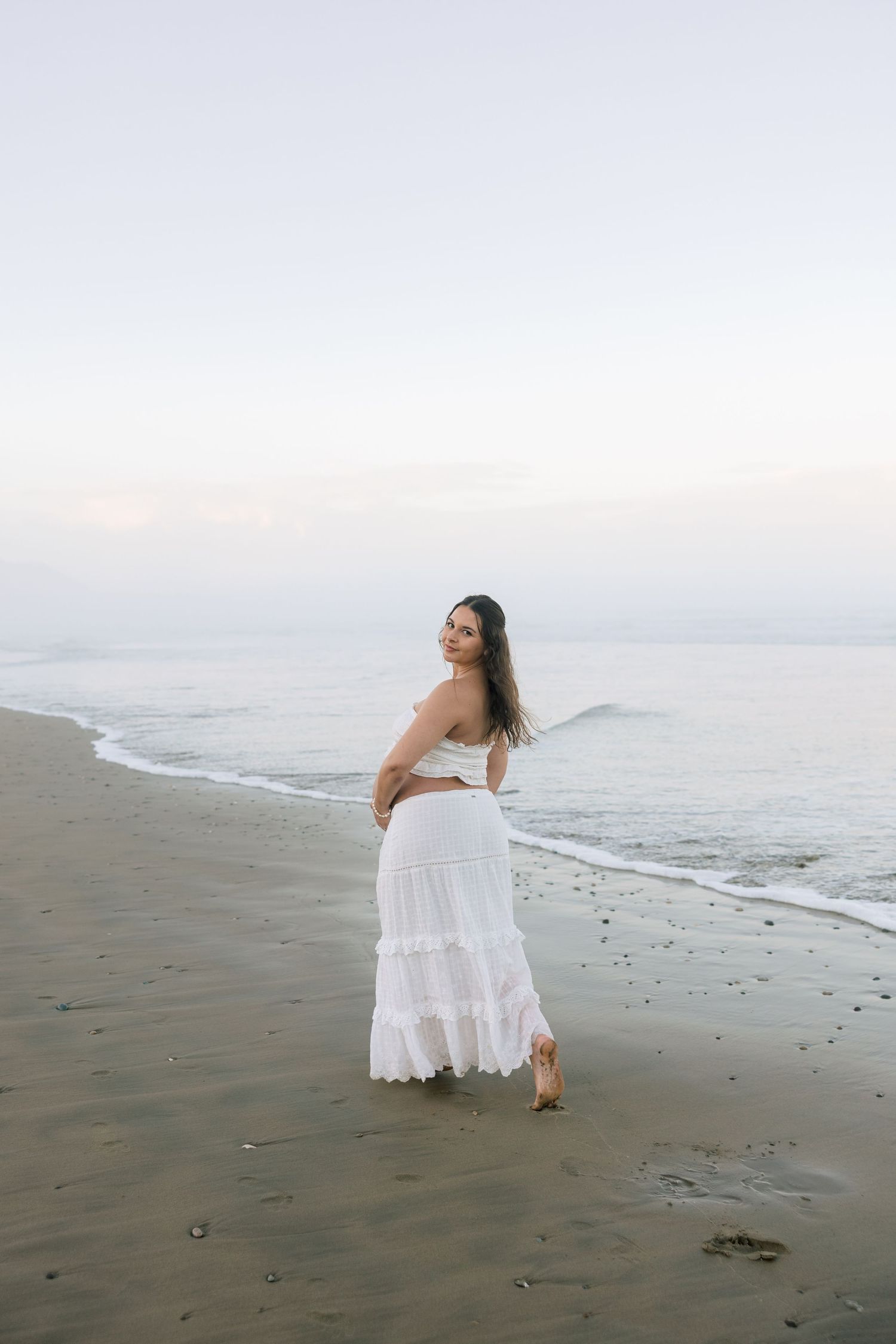 Woman in white strapless top and maxi skirt standing on a sandy beach at dusk with ocean waves.