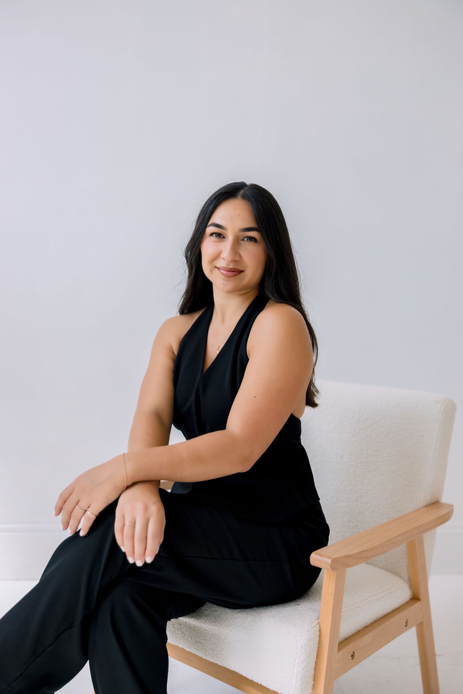 Professional lifestyle portrait series showing person in black dress seated on light wooden chair against white background.