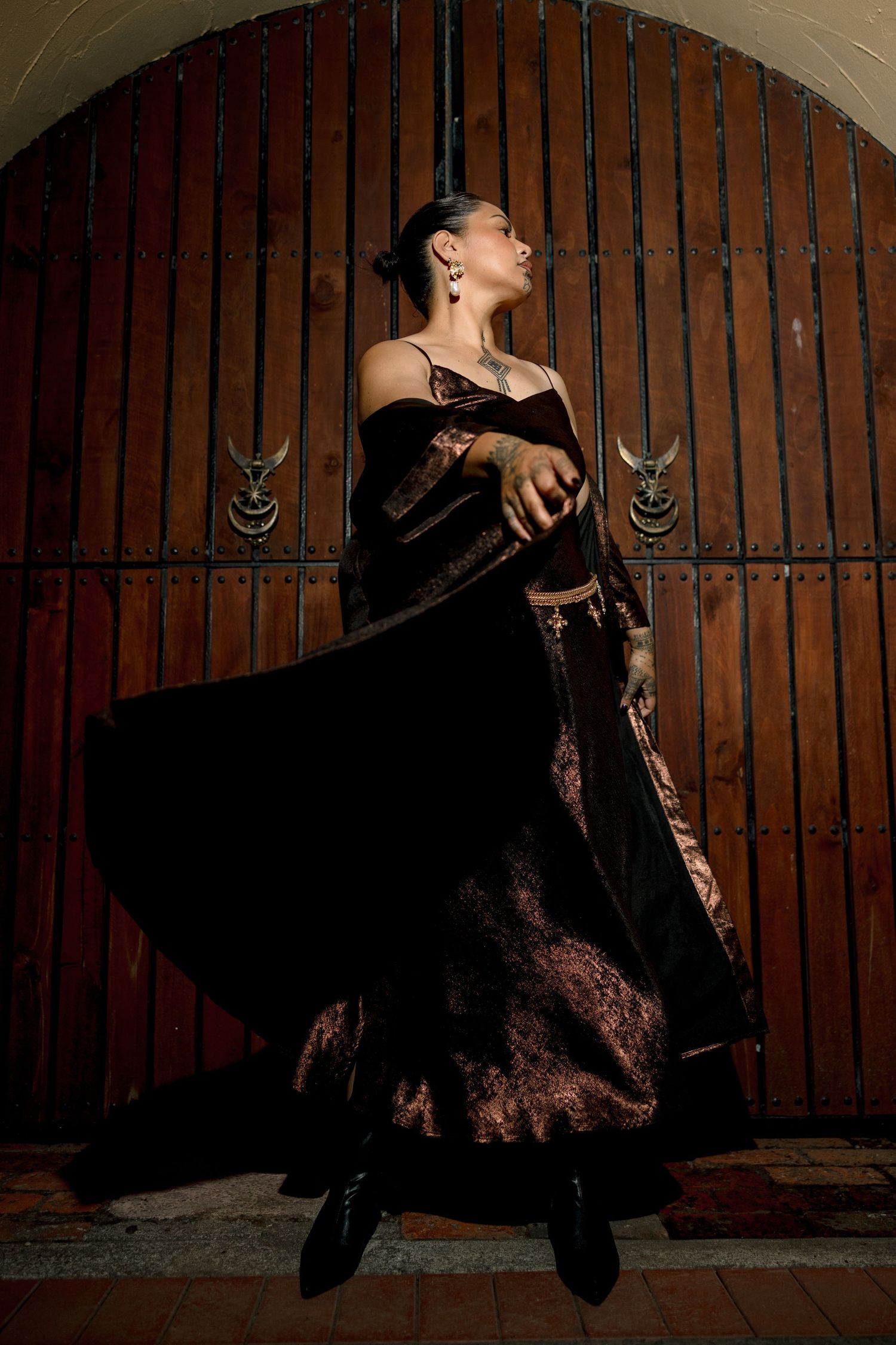 Dramatic fashion portrait of dancer in flowing black dress and boots posing against rustic wooden door with metal accents.