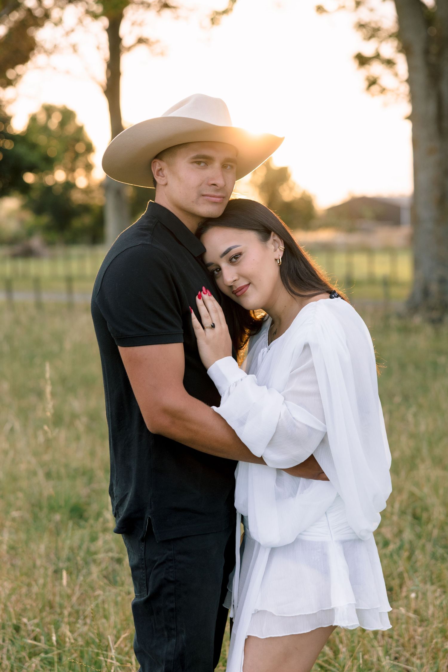A couple embraces in a sunlit field at golden hour with trees silhouetted in the background.