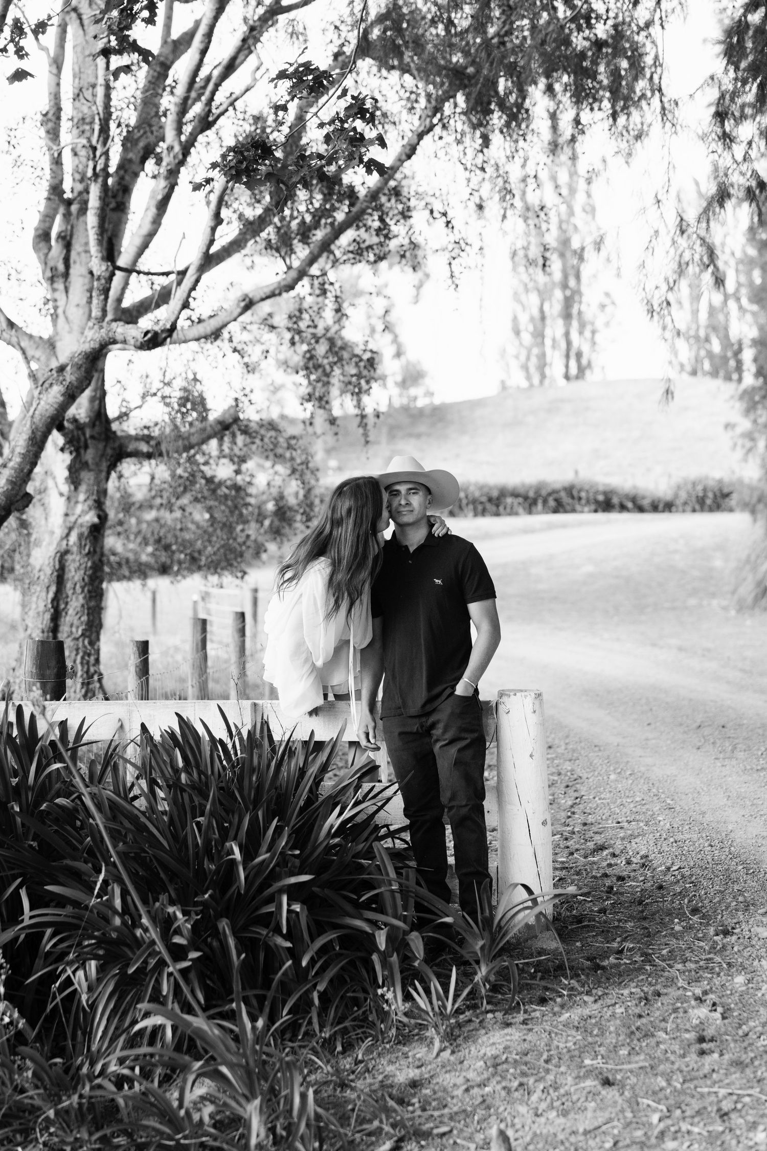 Black and white portrait of a couple posing by a rustic fence with trees in the background.