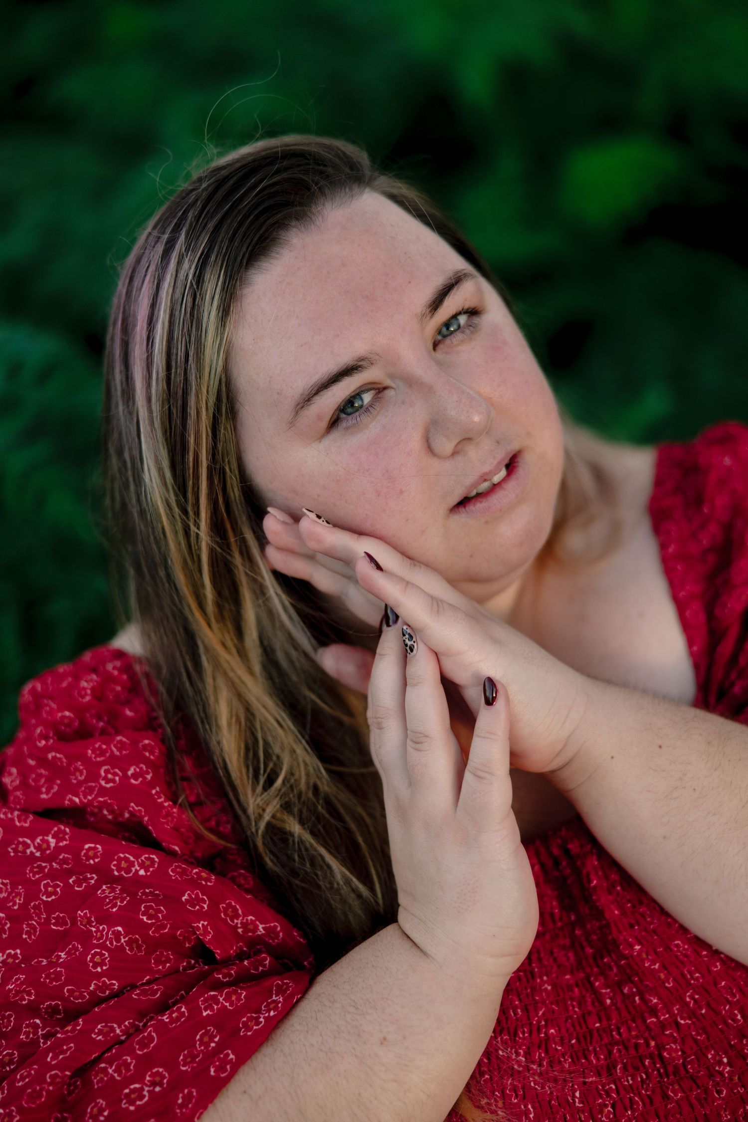 A young woman in a red floral dress rests her hand on her cheek, posing outdoors with green foliage behind her.