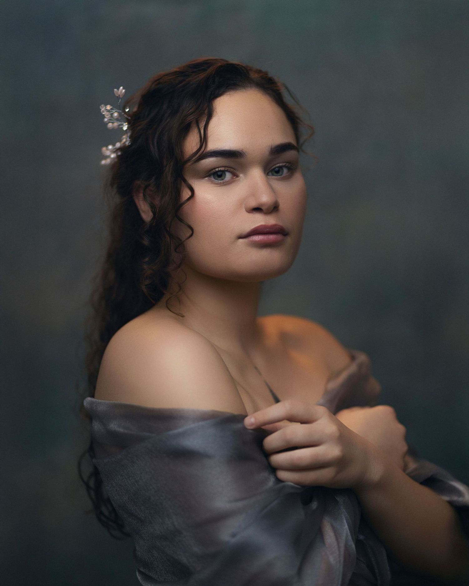 Young woman in off-shoulder gray gown with floral hair accessories poses elegantly against a dark teal studio backdrop.