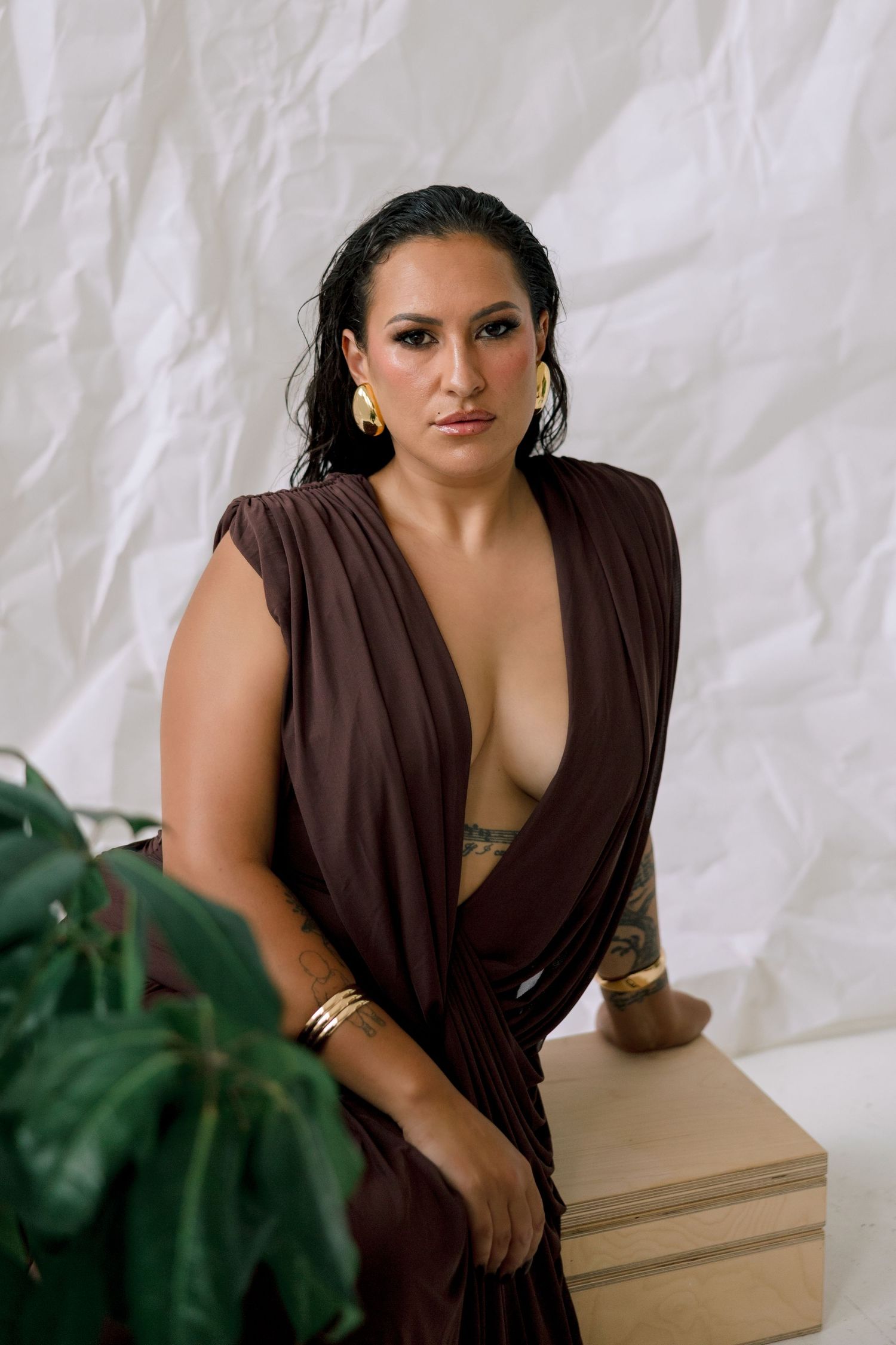 Woman in brown draped dress with gold jewelry sits on wooden box against white backdrop with tropical plant.