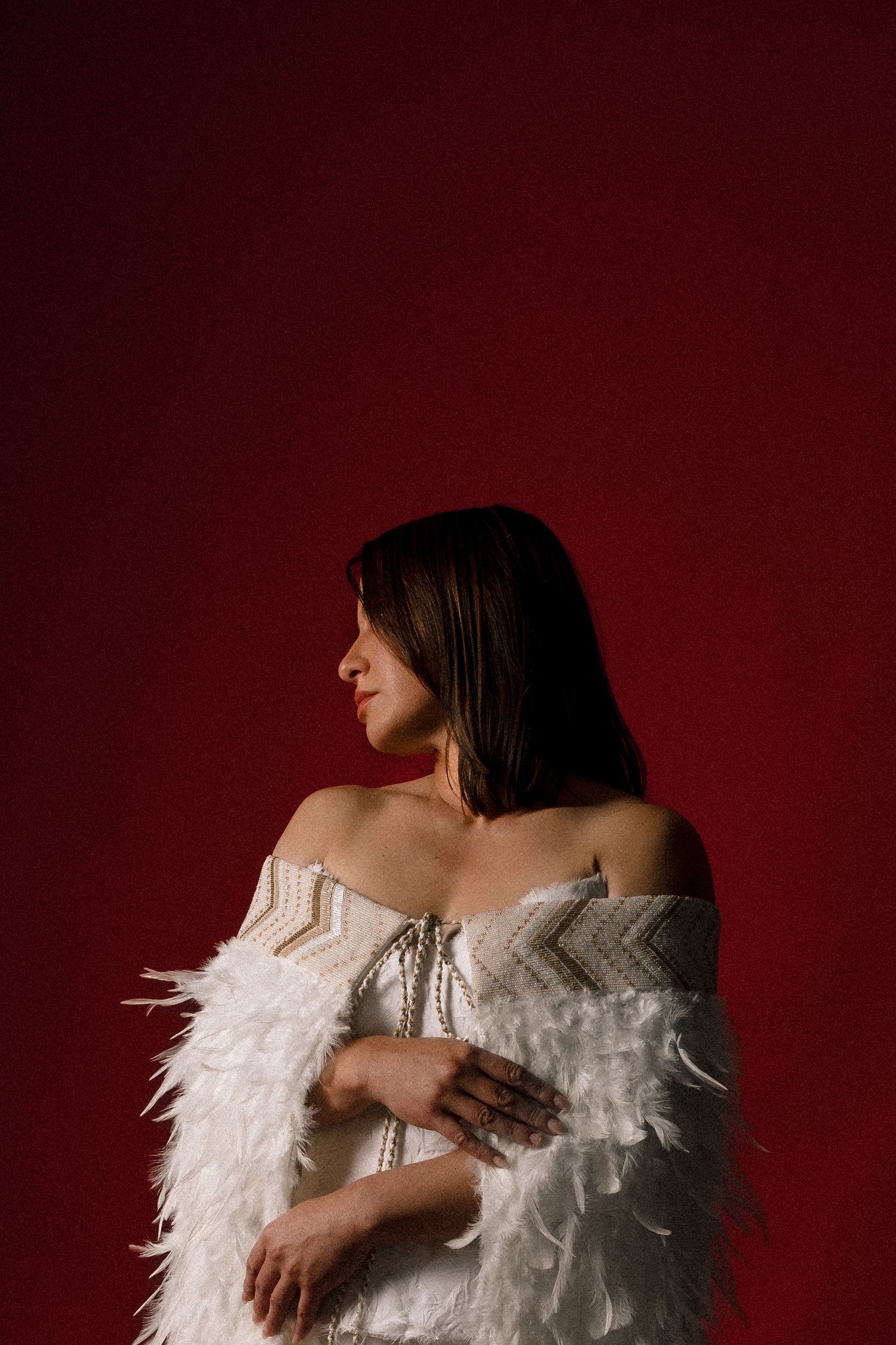 Woman in white feathered off-shoulder gown with beaded details poses against a bold red background.