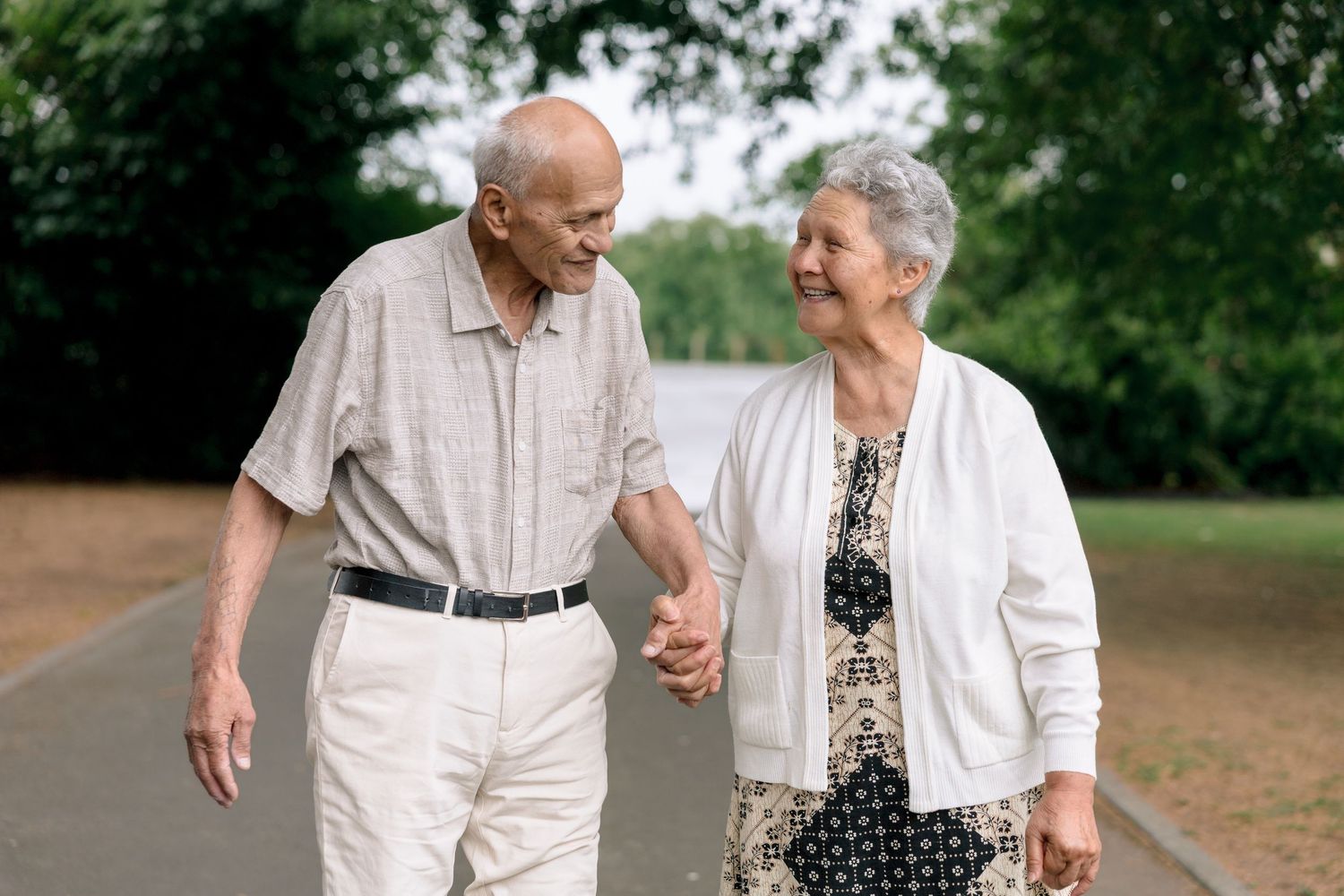 An elderly couple holding hands while taking a walk together on a dirt path surrounded by green trees.