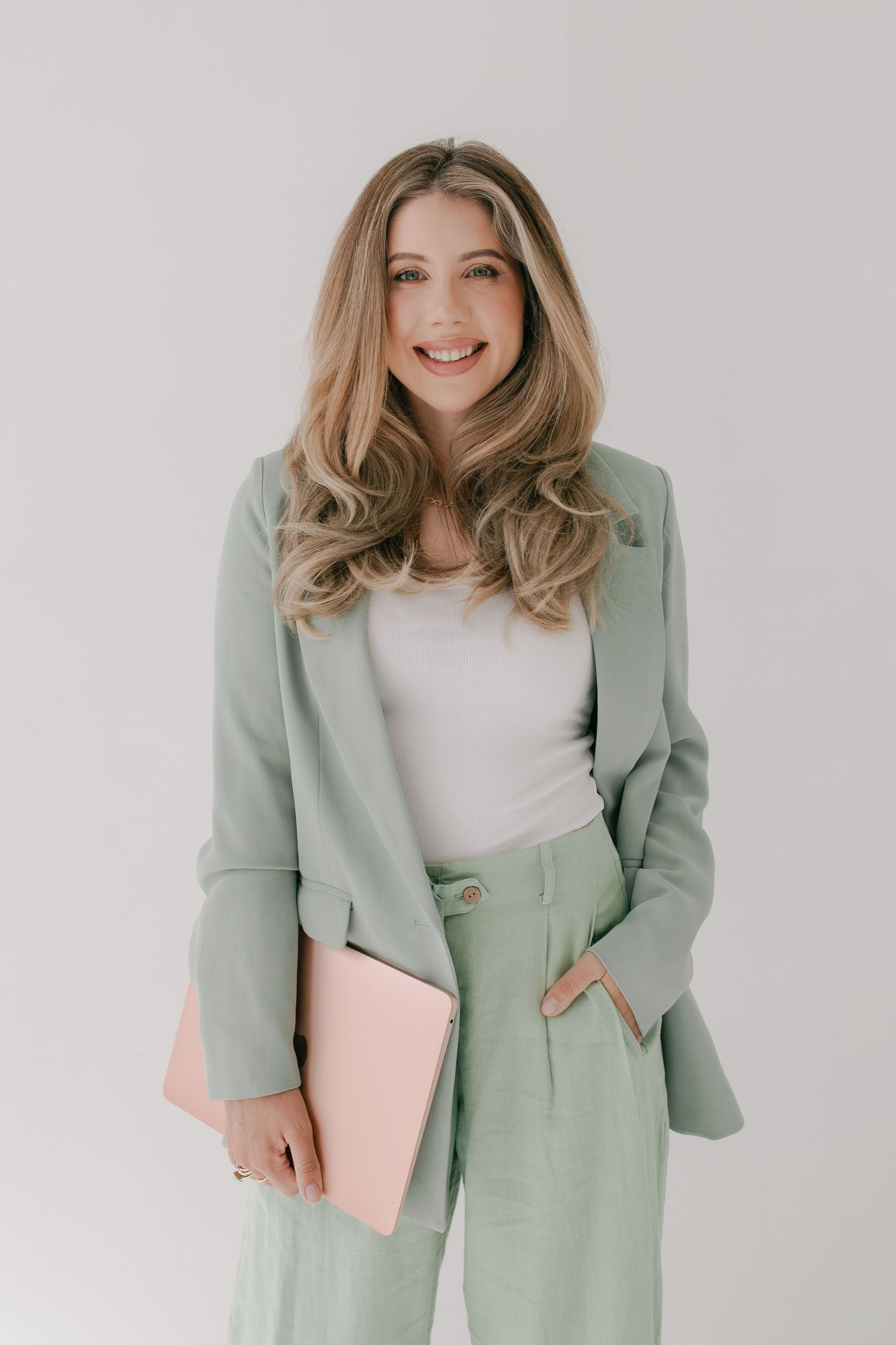 Professional portrait in sage green pantsuit with blush pink laptop case against white background.