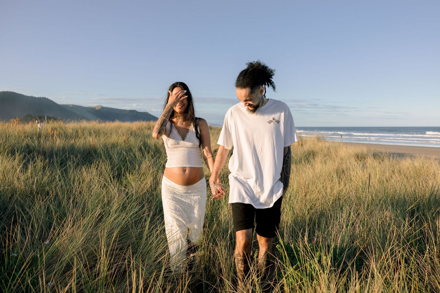 Pregnant couple holding hands walking through tall grass near a beach with mountains in the background.