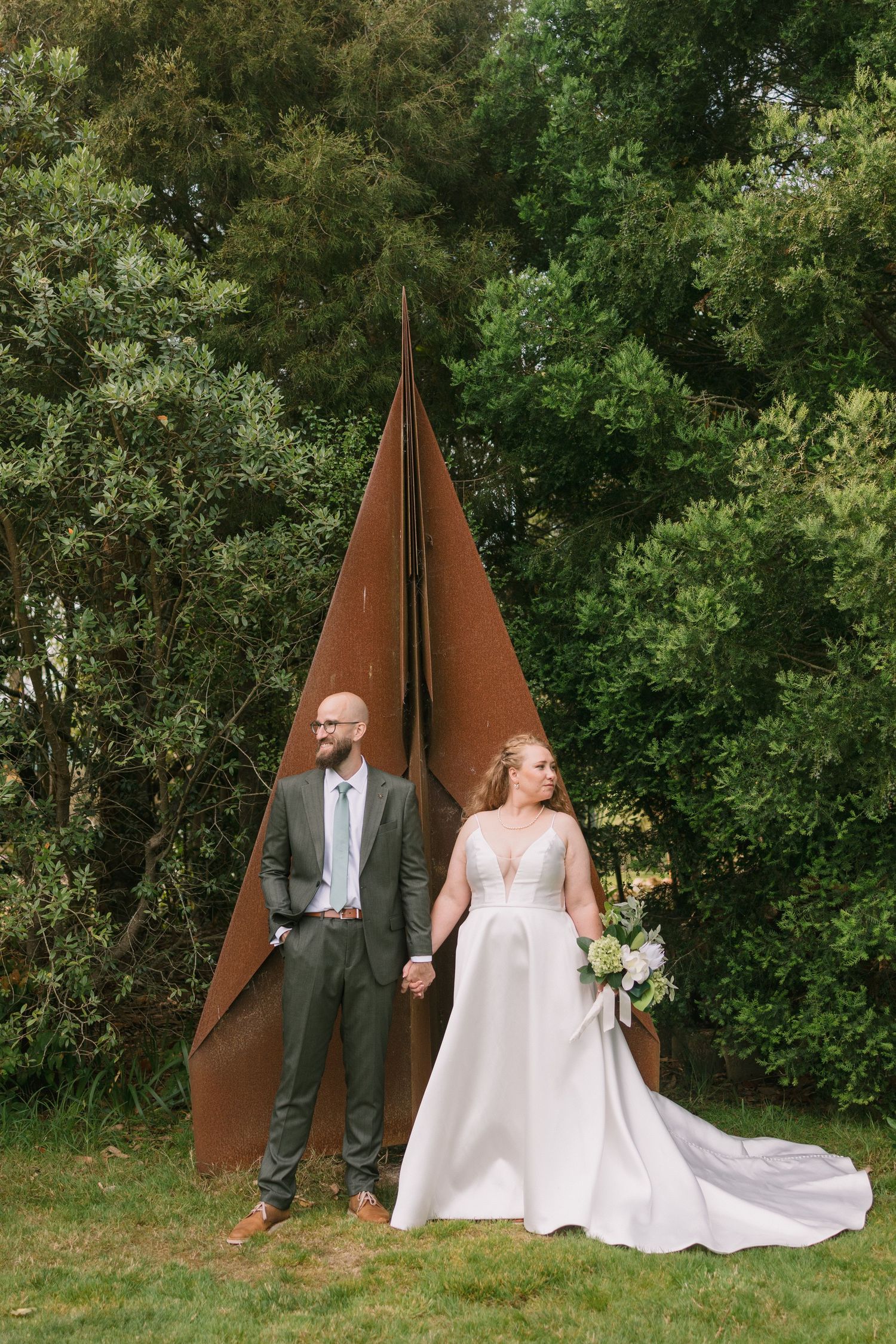 Newlyweds pose in front of a geometric metal sculpture in a garden setting.