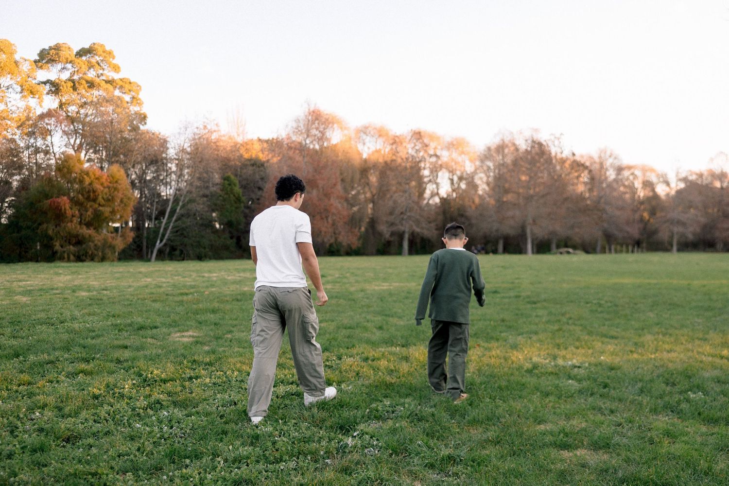 A parent and child walk together across a lush green park field surrounded by autumn trees at sunset.