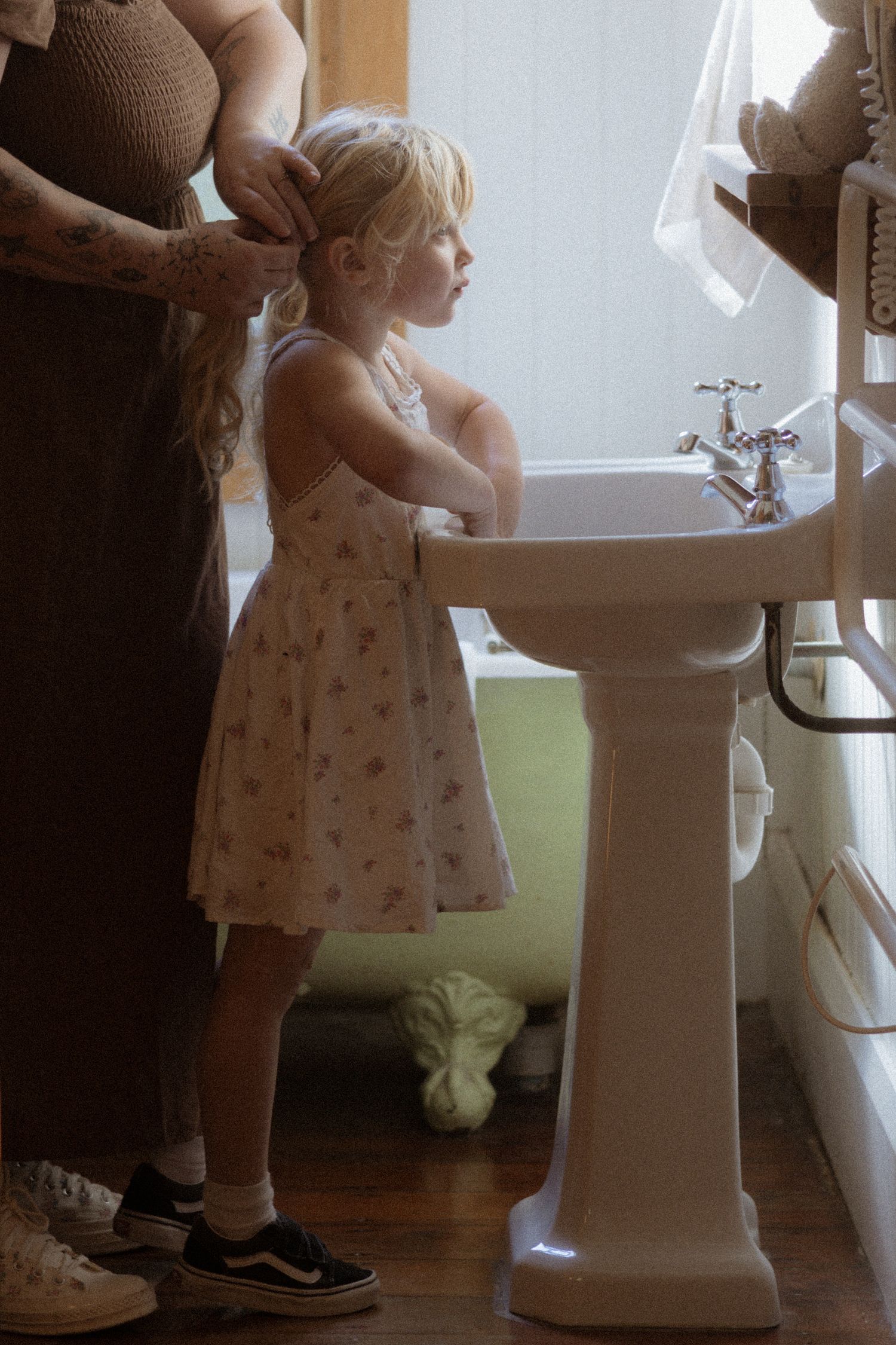 Young child standing at a pedestal sink in a floral dress while getting hair done in a sunlit bathroom.