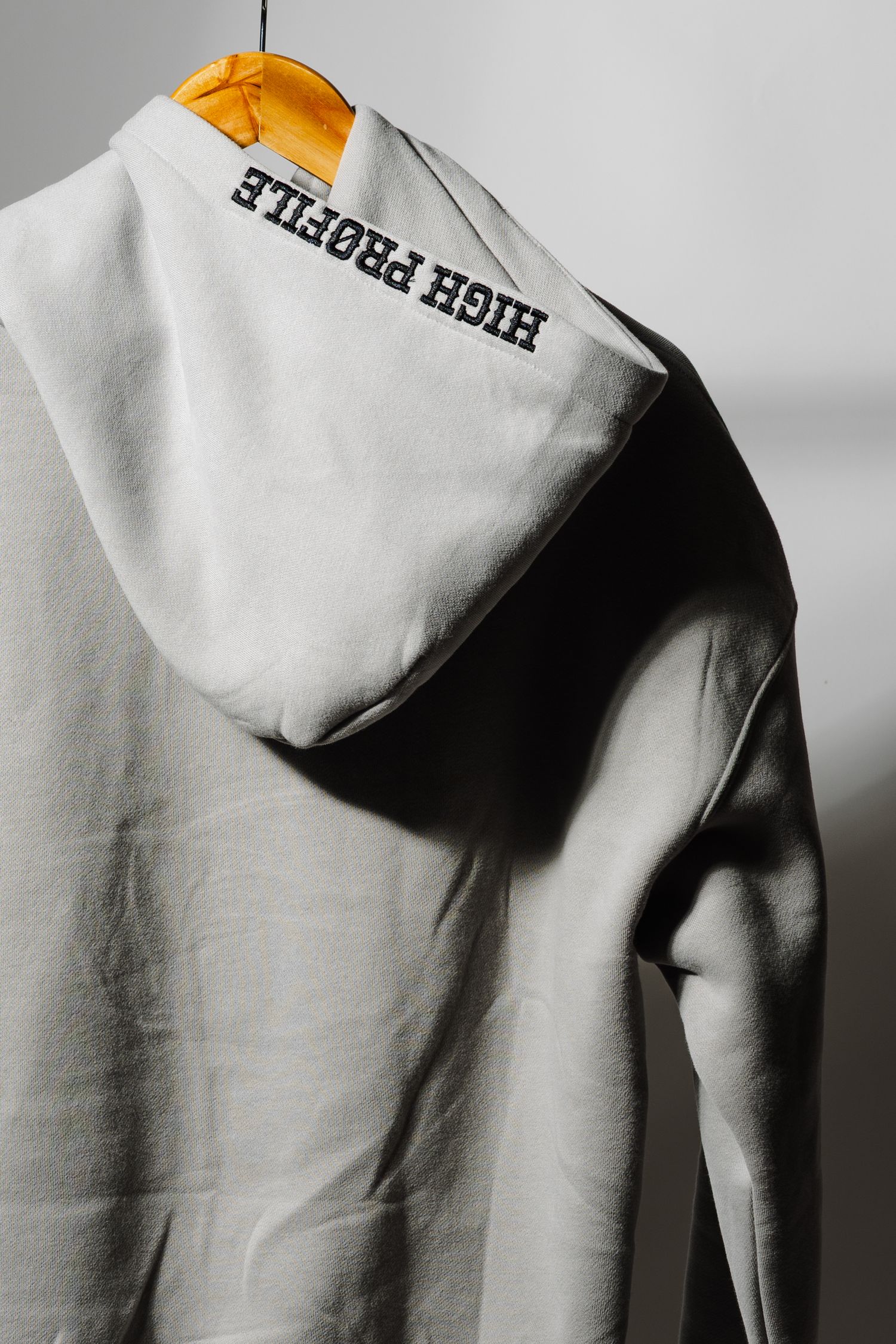 Close-up of light grey hoodie with High Profile text branding on sleeve hanging on yellow hanger.