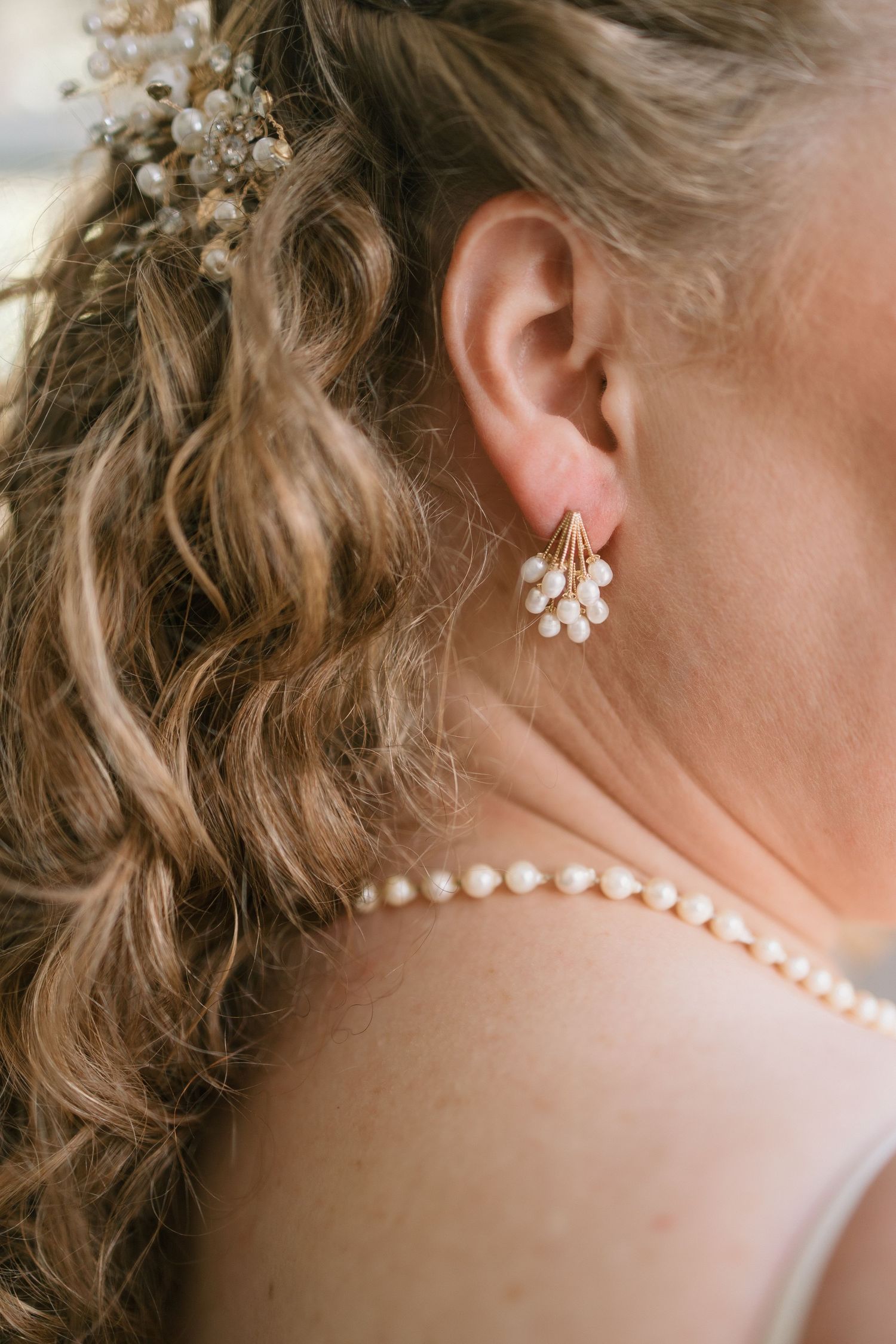 Close-up detail of bridal jewelry showing pearl earrings and necklace with curled blonde hair and floral hair accessories.