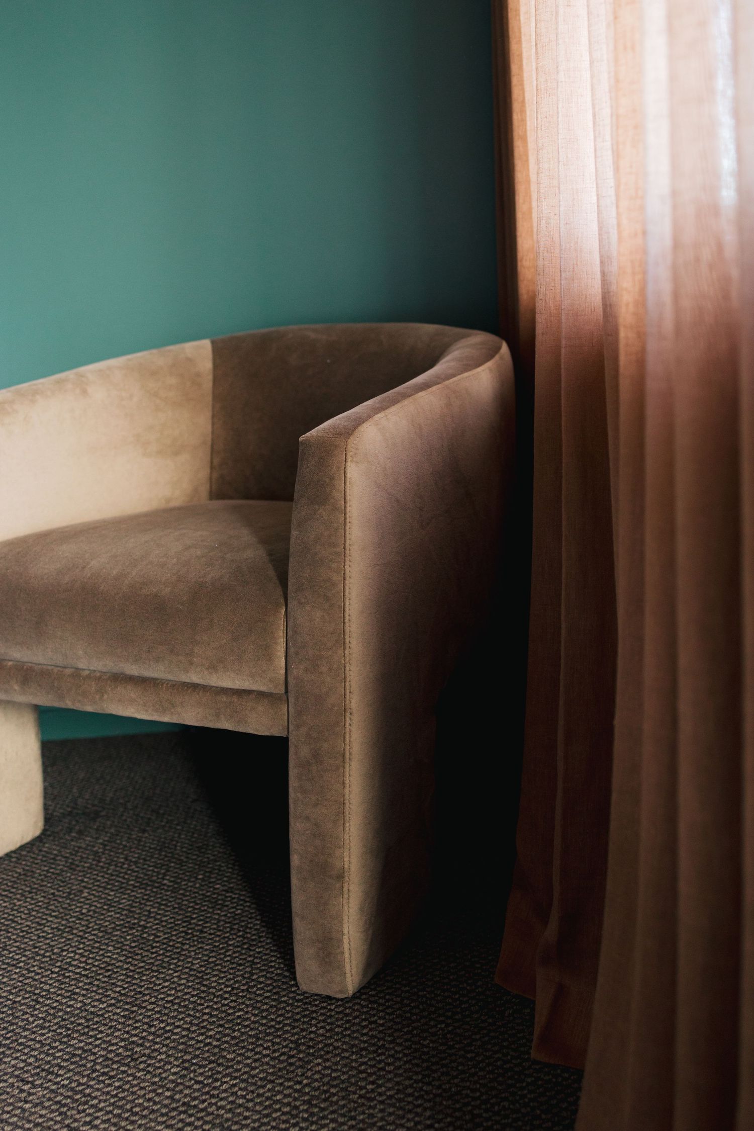 Tan velvet accent chair against dark green wall with flowing curtains.