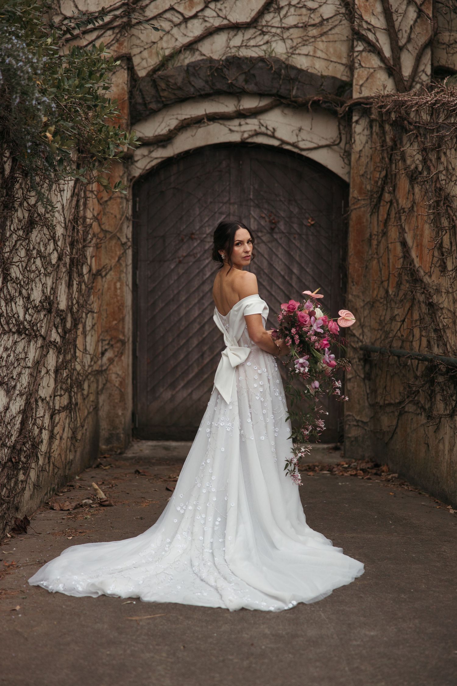 A bride in an elegant off-shoulder lace wedding dress holds a burgundy bouquet against a rustic wooden door backdrop.