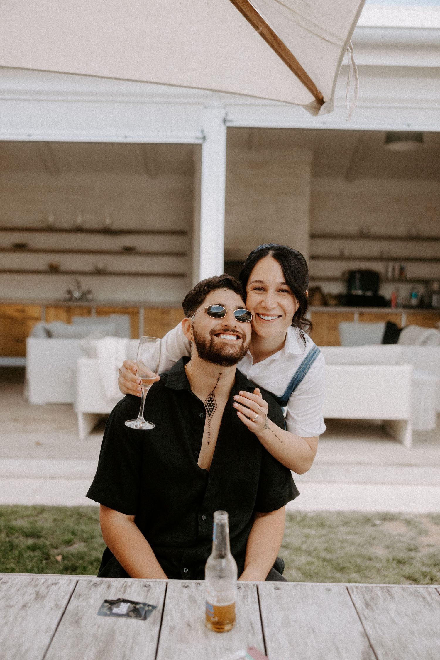 Couple enjoying drinks and laughs together on outdoor wooden deck with white furniture and umbrellas.
